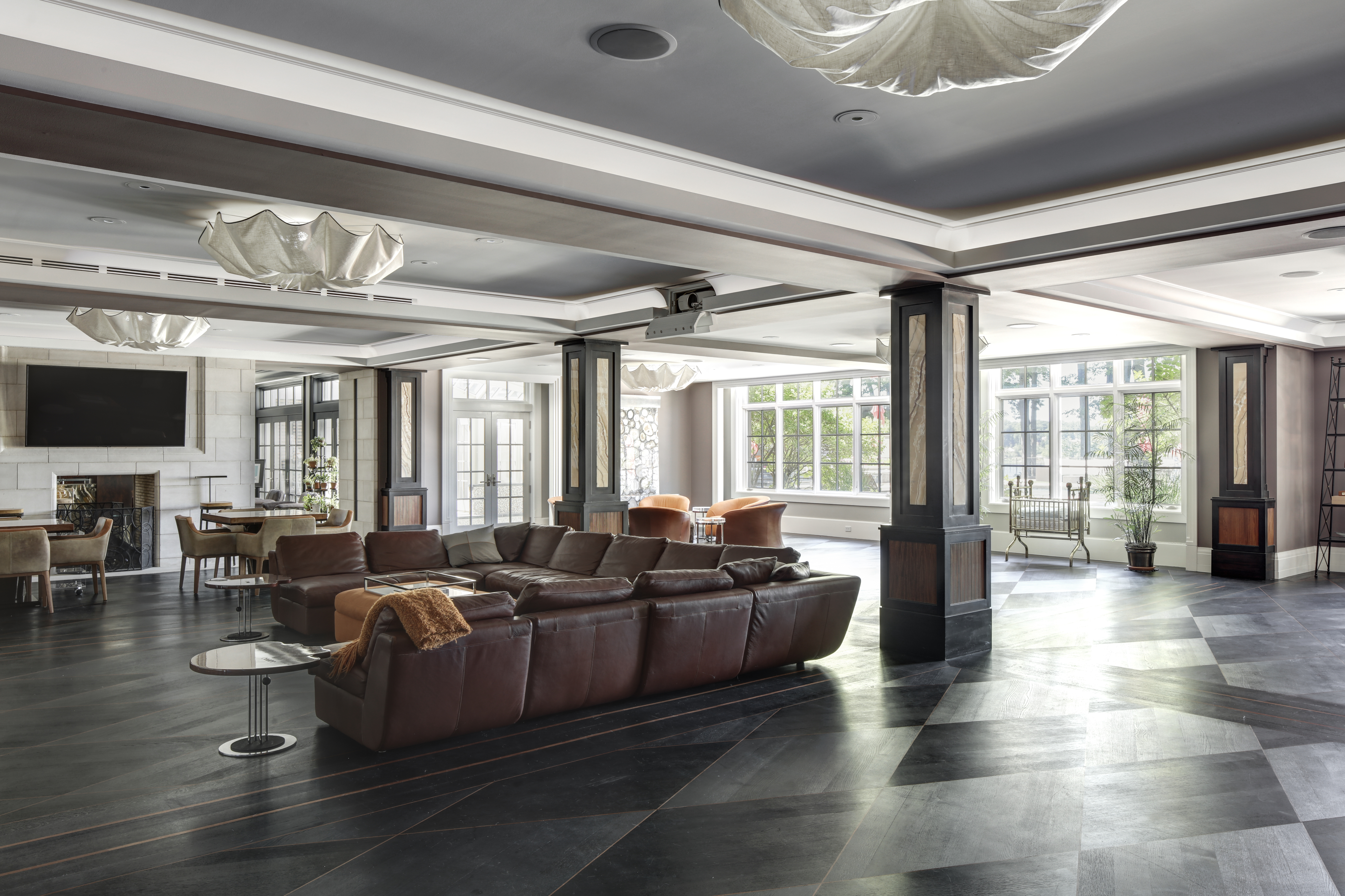 This expansive living room features a large, sectional leather sofa as a focal point, complemented by modern side tables. The room is illuminated by unique, cloud-like pendant lights and natural light streaming through large windows. The dark, patterned flooring adds a touch of sophistication, while the overall design creates a luxurious and inviting atmosphere.