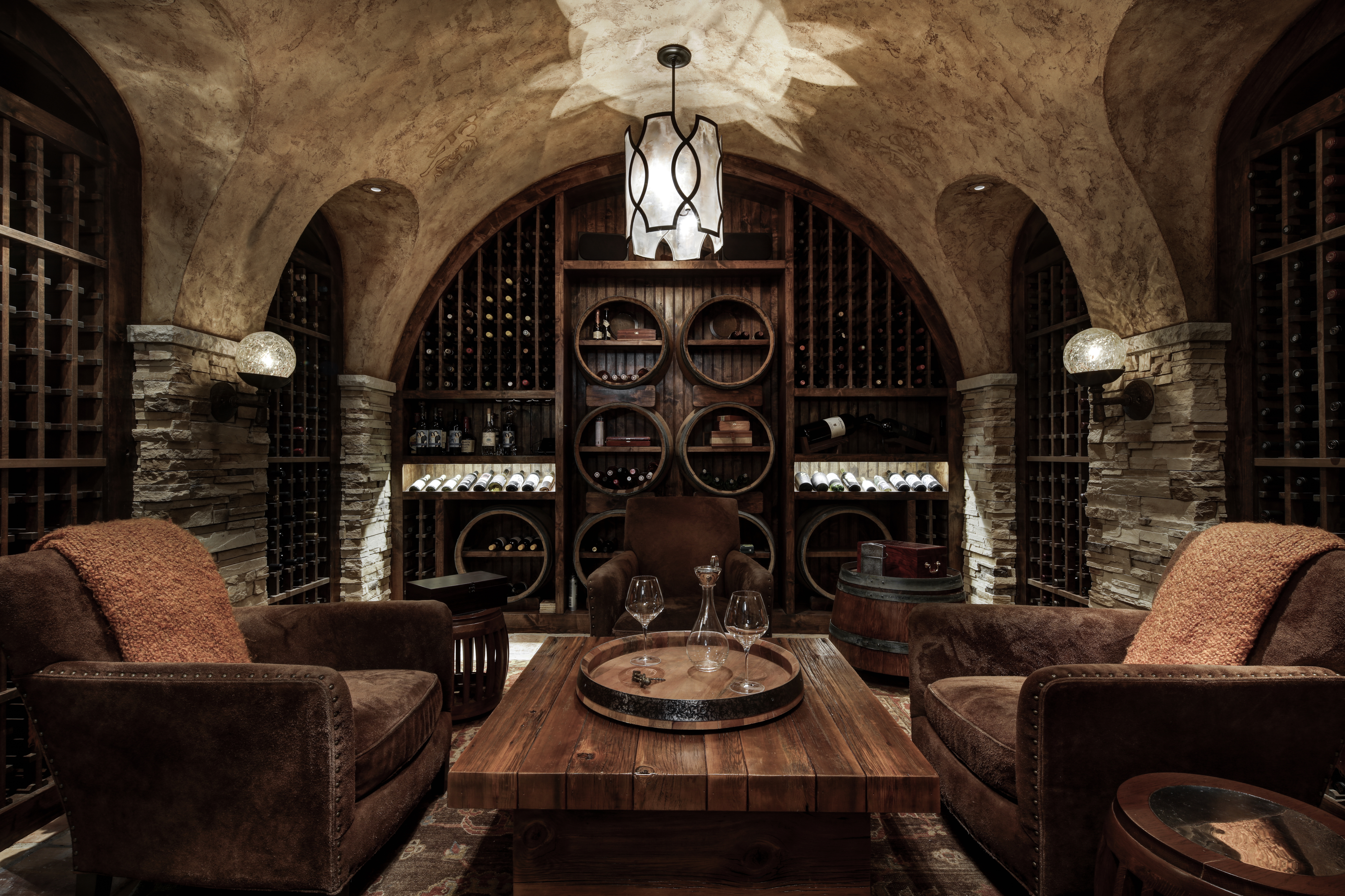 This is an inviting wine cellar featuring a rustic design with stone accents and arched ceilings. The room is well-stocked with wine racks and barrels, and a central wooden table with wine glasses creates a tasting area. Two comfortable armchairs flank the table, suggesting a cozy space for relaxation and enjoyment.