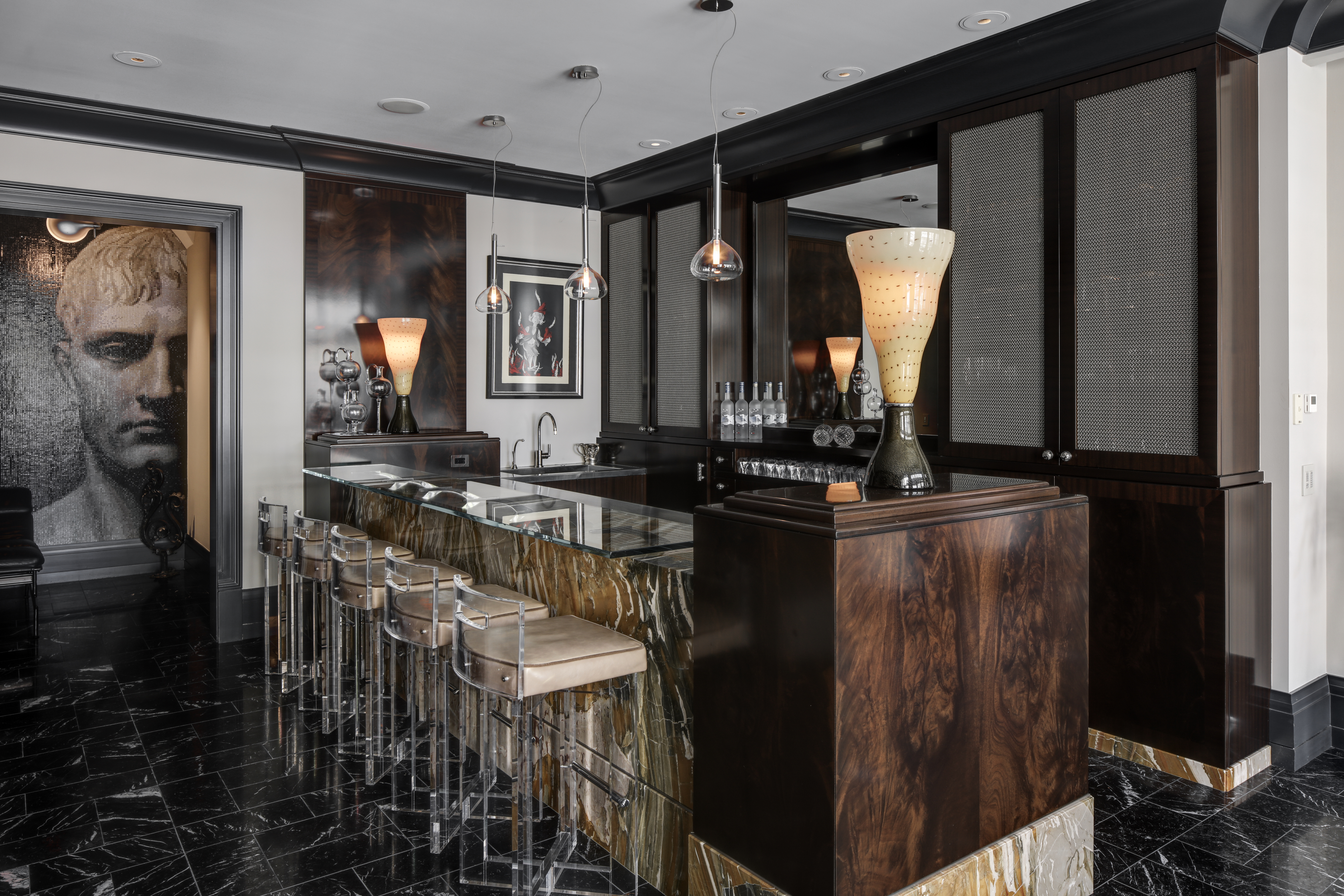 This interior shot showcases a sophisticated wine cellar featuring a marble-topped bar with clear acrylic stools. The room is adorned with dark wood cabinetry, decorative lighting, and a striking mosaic portrait, creating a luxurious and inviting atmosphere. The black marble flooring adds to the room's elegance.