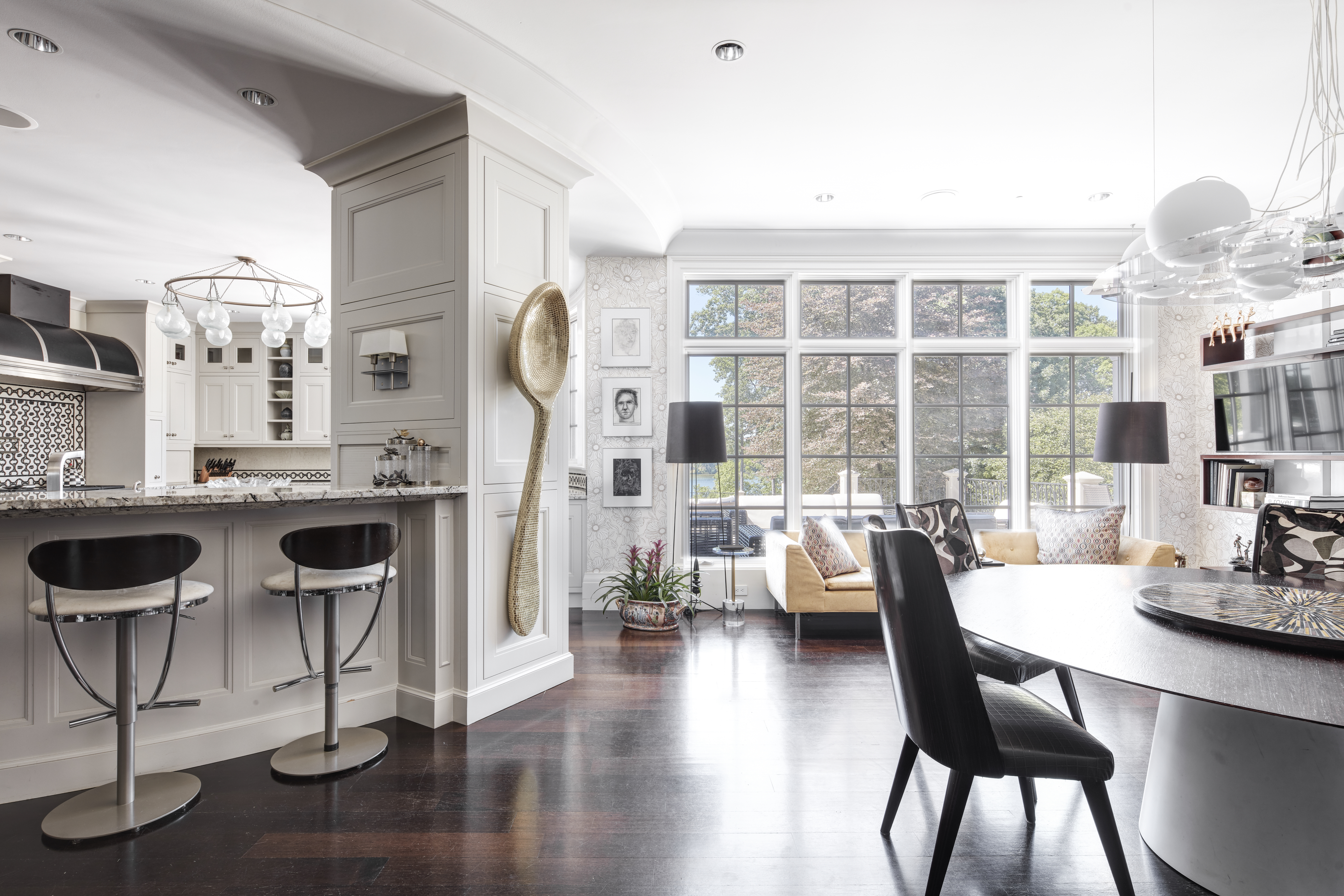 This is an interior shot showcasing an open-concept living and dining area. The space features dark hardwood floors, a modern dining table with chairs, and a comfortable seating area with a large window providing natural light. A kitchen bar with stools is visible in the background, and a decorative oversized spoon adorns a wall, adding a unique artistic touch.