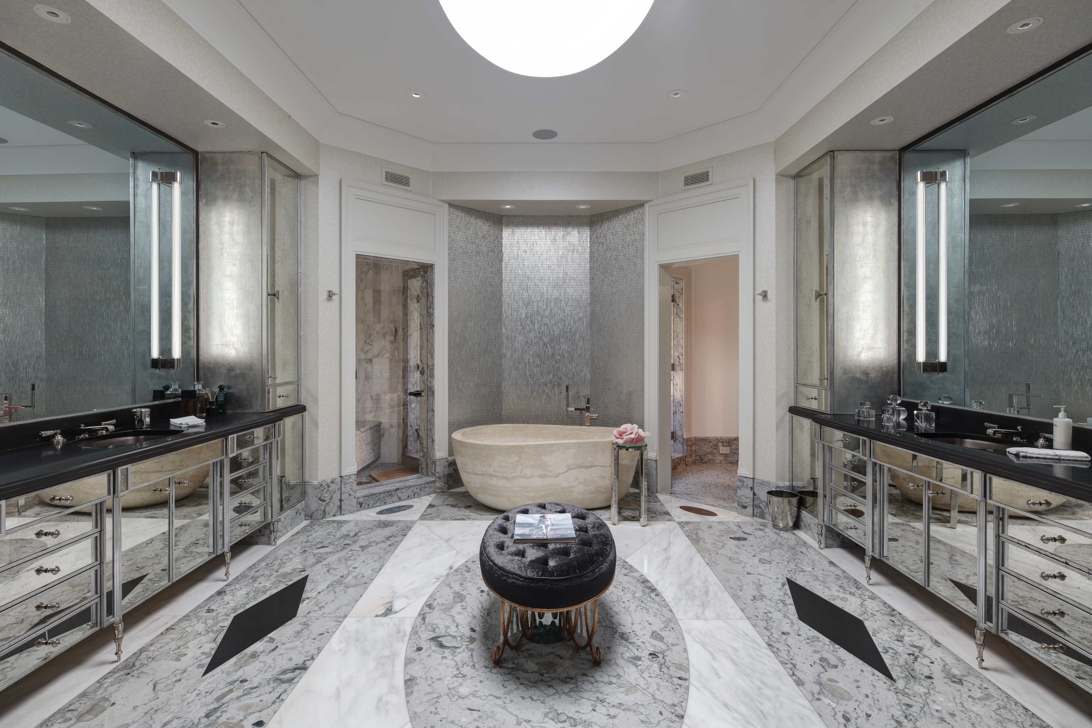 This is a luxurious primary bathroom featuring a freestanding stone bathtub as a focal point, flanked by doorways leading to other areas. The room boasts dual mirrored vanities with dark countertops, and the floor is a striking combination of white and gray marble with black accents. A round, tufted ottoman sits in the center of the room, adding a touch of elegance.
