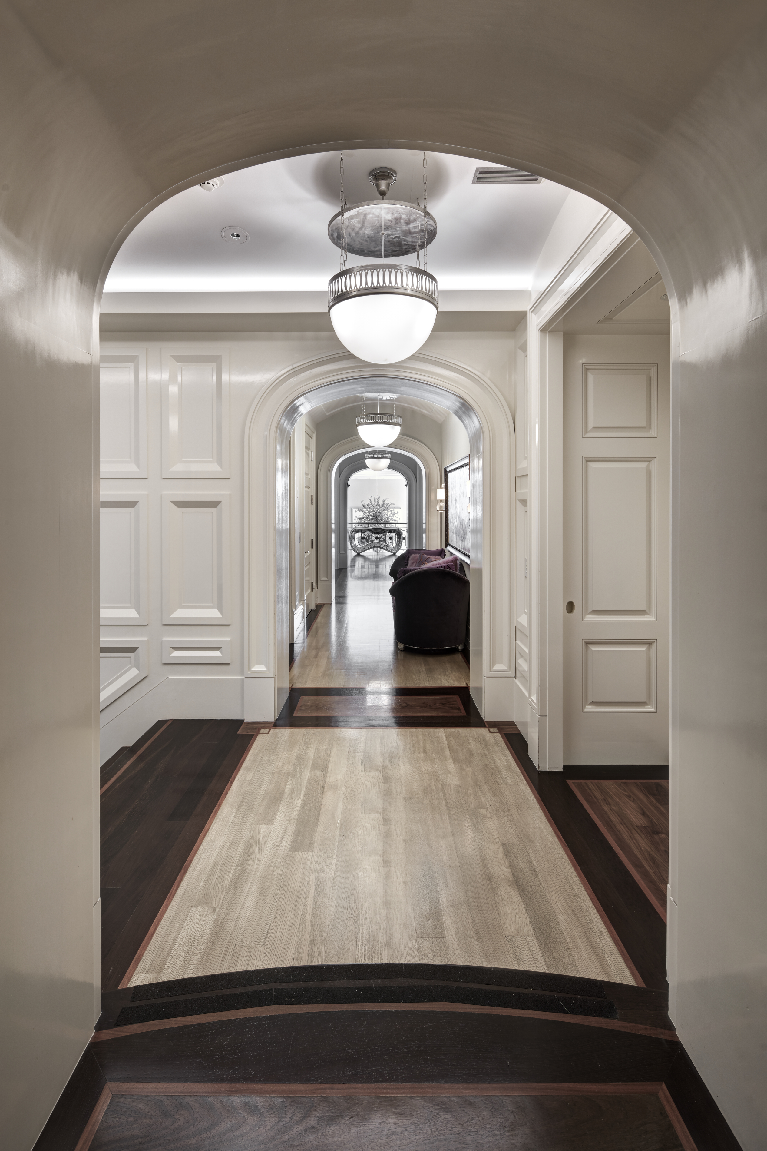 This is an interior shot of a luxurious hallway featuring a series of arched doorways that create a sense of depth and grandeur. The hallway has a unique floor design with a combination of light and dark wood, complemented by white paneled walls and elegant pendant lighting. The perspective emphasizes the length of the hallway, inviting viewers to imagine walking through the space.