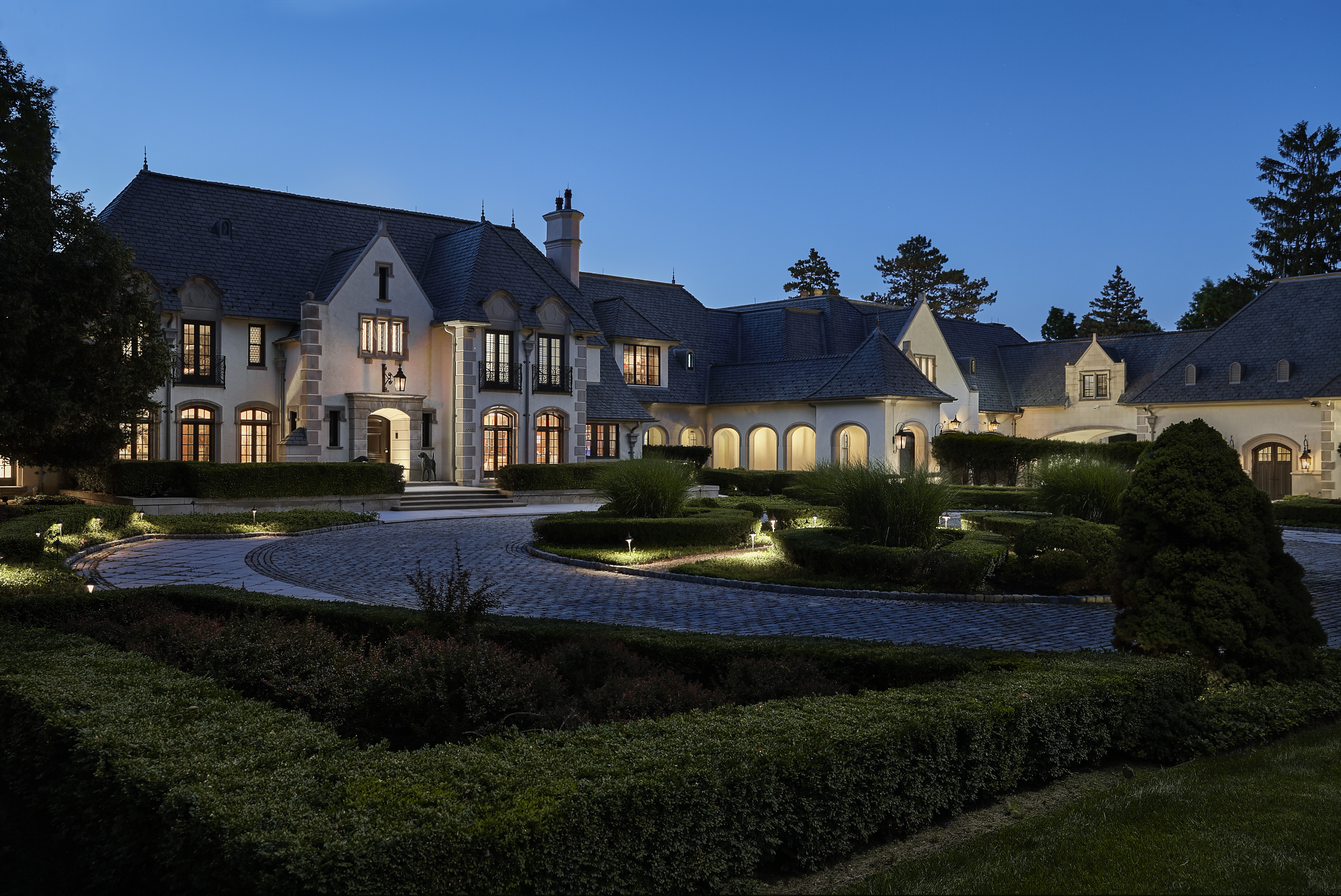 This is a stunning front view of a large, luxurious estate at dusk. The architecture features a blend of classic and modern elements, with a grand entrance, multiple windows with warm interior lighting, and a meticulously landscaped circular driveway and garden. The overall impression is one of elegance, privacy, and high-end living.
