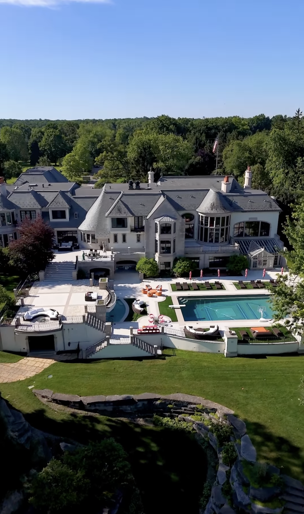 This aerial view showcases a grand estate with a sprawling layout, featuring a large swimming pool, multiple patios, and meticulously landscaped grounds. The architecture is a blend of traditional and modern elements, with a gray roof, light-colored exterior, and numerous windows offering ample natural light. The property is surrounded by lush greenery, providing privacy and a sense of seclusion.