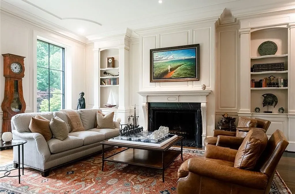 This elegant, formal living room features traditional architectural details, including wall moldings and built-in shelving flanking a black marble fireplace. The space is comfortably furnished with a neutral-toned sofa and two rich leather armchairs, all centered around a coffee table displaying a large chess set. An ornate oriental rug and a classic grandfather clock complete the sophisticated, warm ambiance of the room.