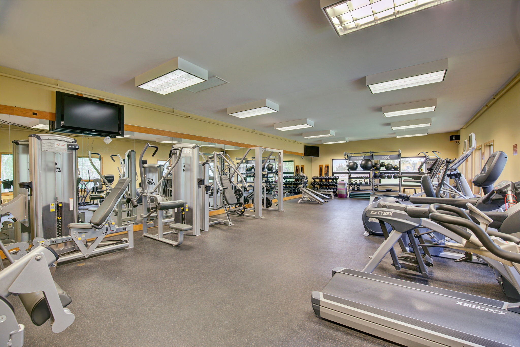 This spacious and well-equipped fitness center features a variety of professional-grade strength training machines and treadmills arranged on a durable rubberized floor. The room is brightly lit by multiple large ceiling lights and includes a mirrored wall that enhances the perception of space while providing feedback during workouts. This facility offers essential amenities for a convenient and comprehensive personal fitness routine.