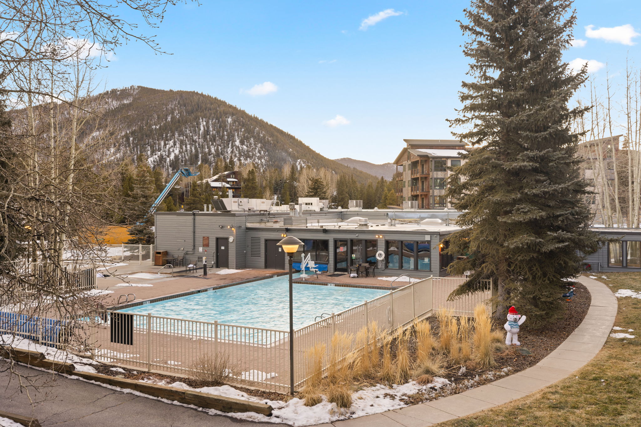This outdoor space features a well-maintained, fenced-in pool that provides a relaxing amenity for residents. The area is set against a scenic backdrop of mountains and mature evergreen trees, offering a tranquil environment. A paved path, lawn, and a playful snowman statue add character to the setting, making it an inviting space for leisure.