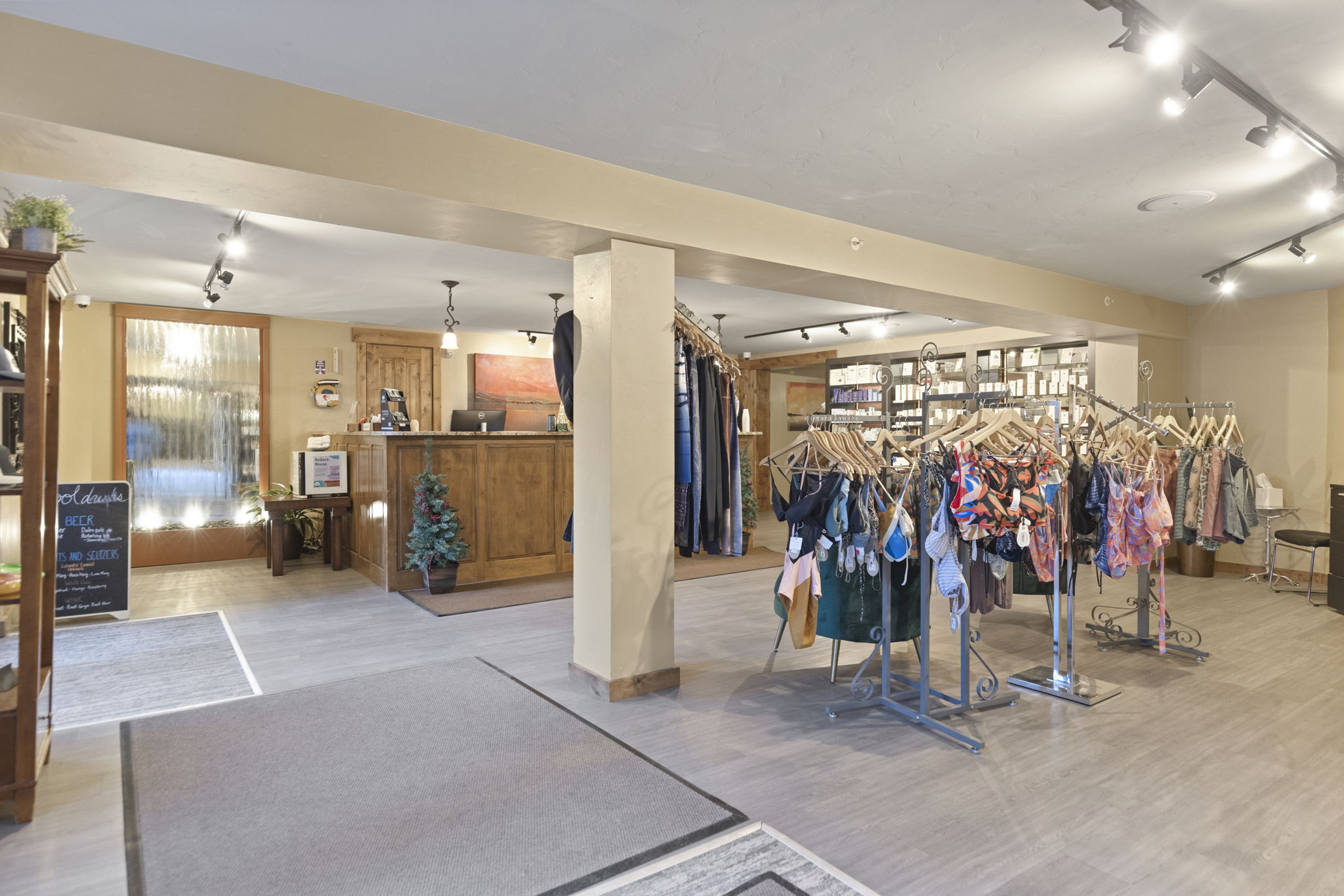 This image shows the interior of a commercial or community activity space, featuring a boutique retail area displaying swimwear and apparel on racks. The space includes a wooden reception desk in the background, light wood-toned flooring, and warm, recessed cove and track lighting. The environment appears well-maintained and serves as both a central service hub and a shopping area for guests.