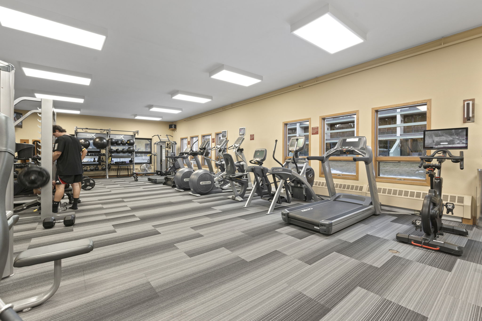 This clean and functional fitness center features a variety of cardio equipment, including treadmills, stationary bikes, and elliptical machines arranged along a row of windows. The space includes a designated weight training area with a rack of medicine balls and benches, all set atop a durable, modern gray-toned patterned carpet. The gym provides a well-lit and organized environment, making it an appealing amenity for residents.
