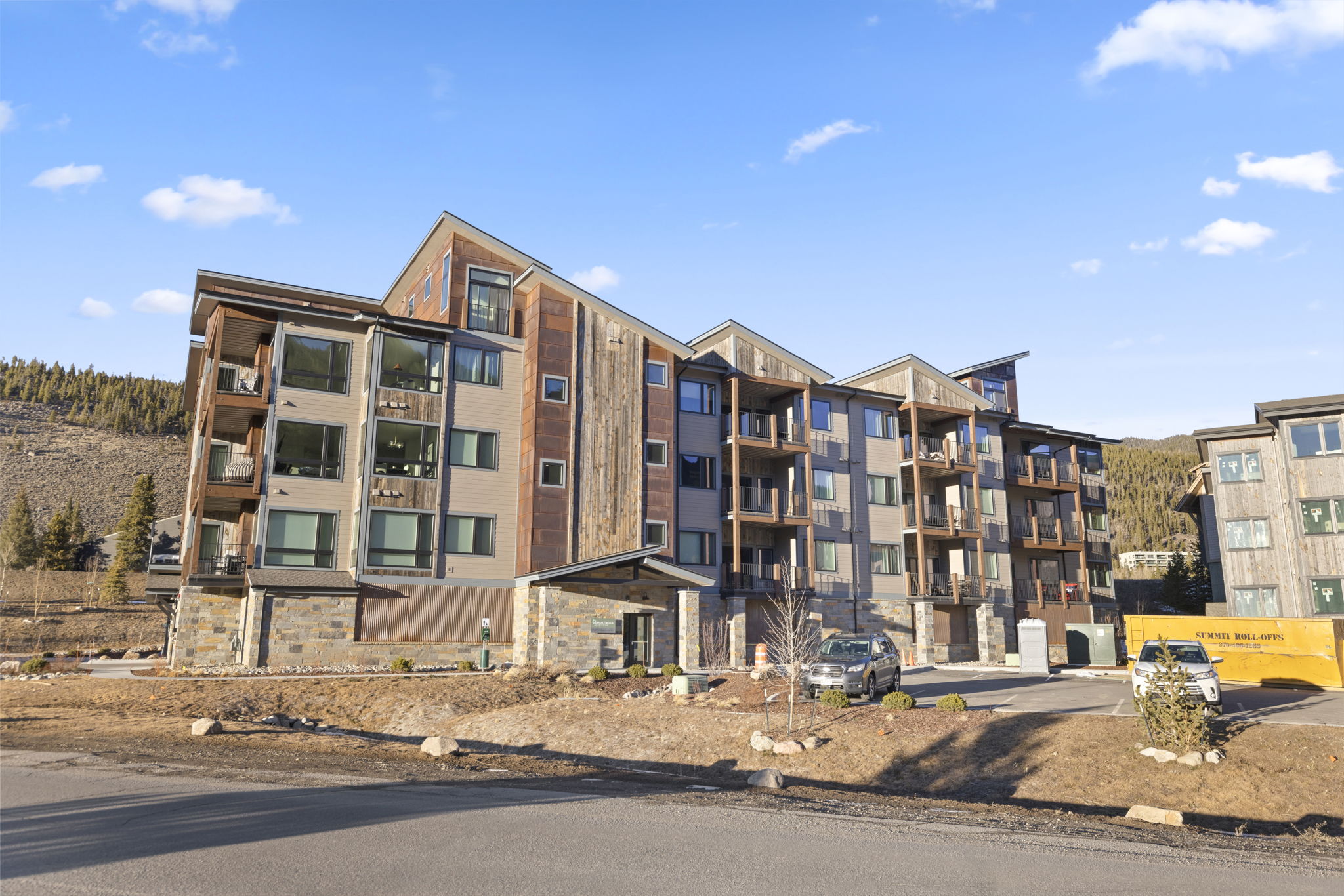 This modern multi-story residential building features a rugged, contemporary aesthetic with a blend of stone masonry, wood siding, and metal accent panels. The complex includes private balconies and large windows that provide abundant natural light, set against a backdrop of mountainous terrain. Its design strikes a balance between mountain-inspired architecture and professional, clean lines suitable for a contemporary condominium or apartment complex.