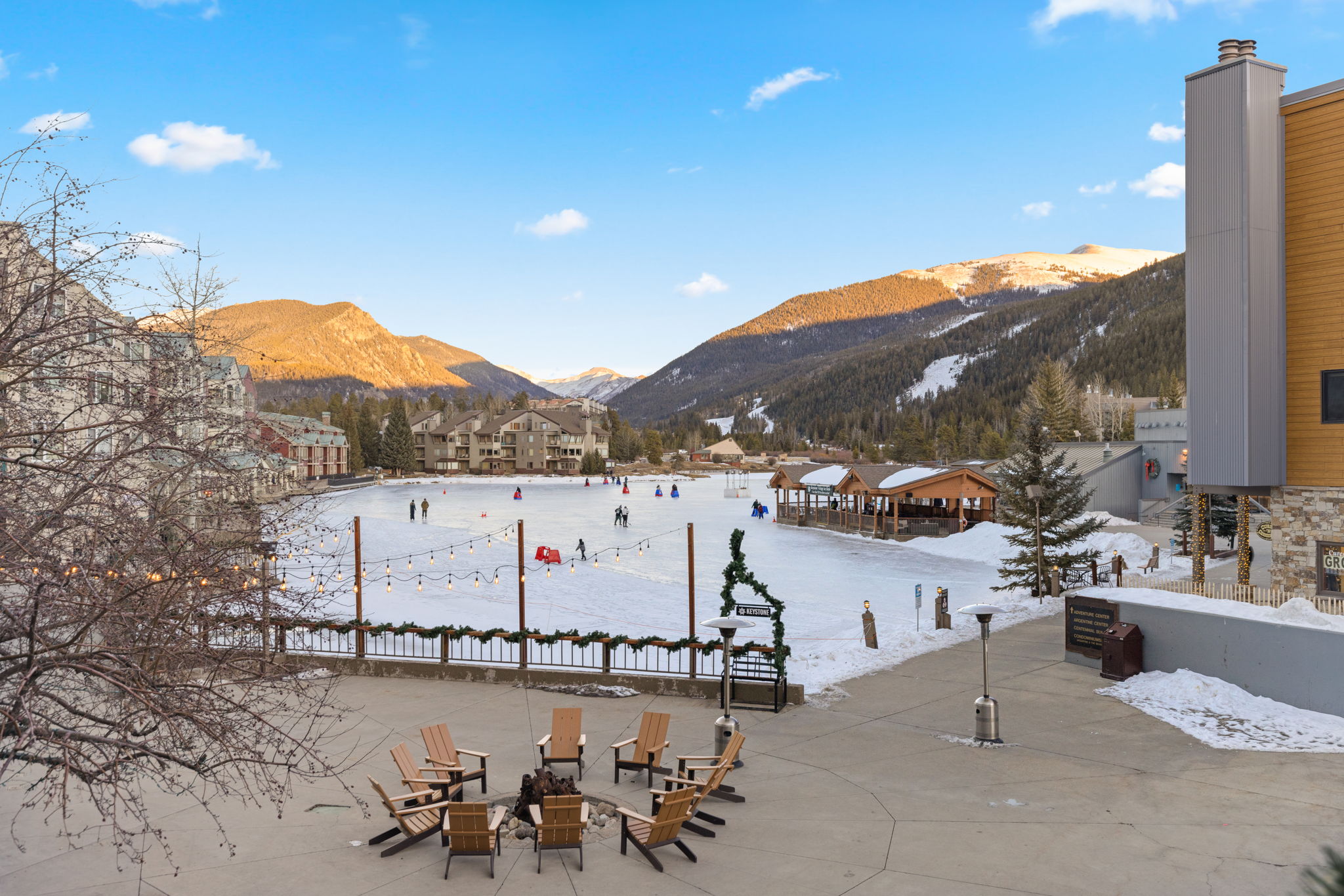 This image showcases a scenic outdoor communal plaza featuring a central fire pit area surrounded by wooden Adirondack chairs. The plaza overlooks a picturesque ice skating rink set against a backdrop of majestic snow-capped mountains and evergreen trees, creating an inviting recreational space. The scene captures a tranquil winter atmosphere, highlighting the desirable lifestyle and amenities offered by the resort-style property.