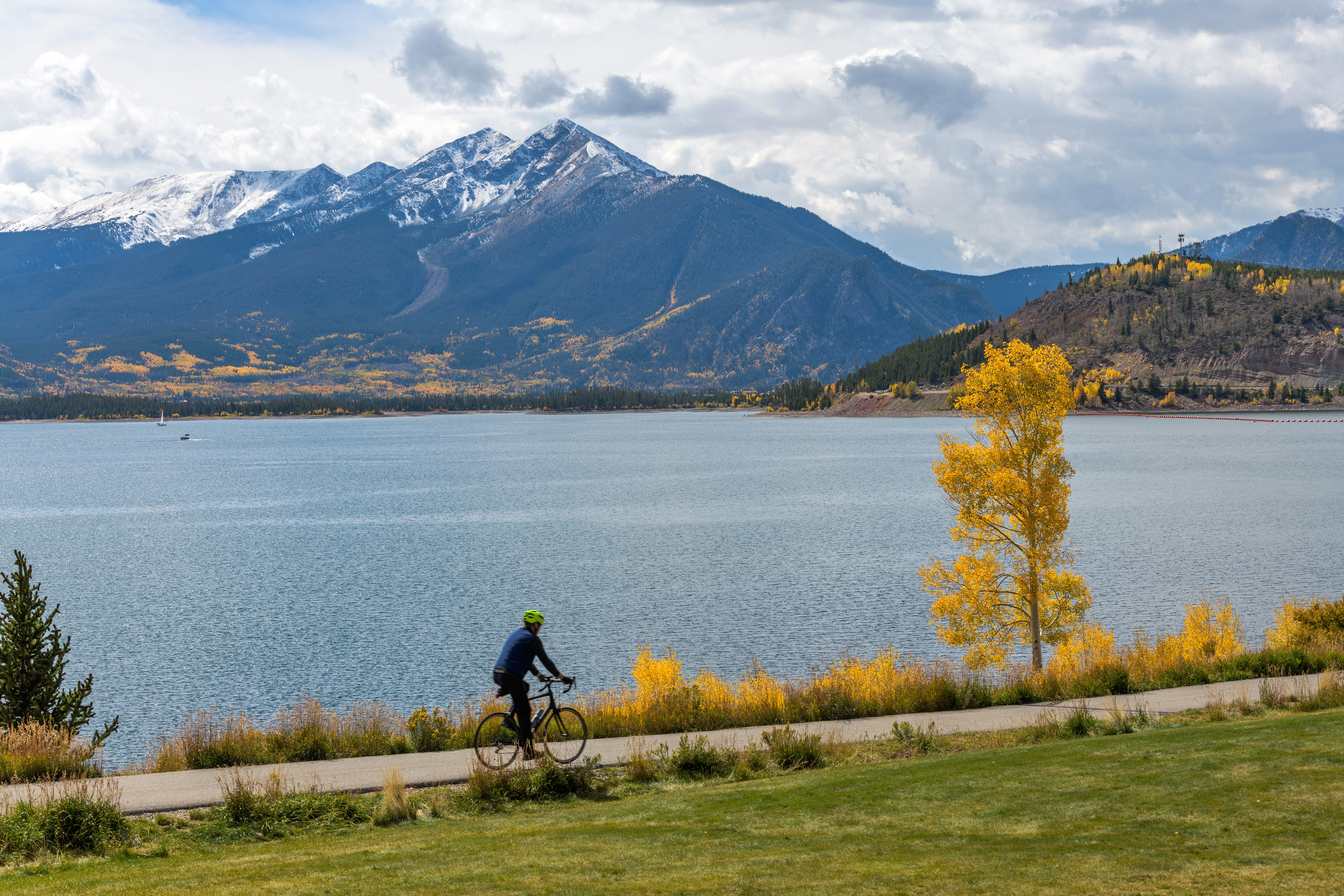 This scenic image captures a paved cycling and walking path overlooking a large mountain lake, showcasing an ideal location for outdoor recreation. The landscape is characterized by vibrant autumn foliage, snow-capped mountain peaks, and a serene waterfront setting. The area provides an attractive, nature-oriented lifestyle feature that is highly desirable for nearby real estate.