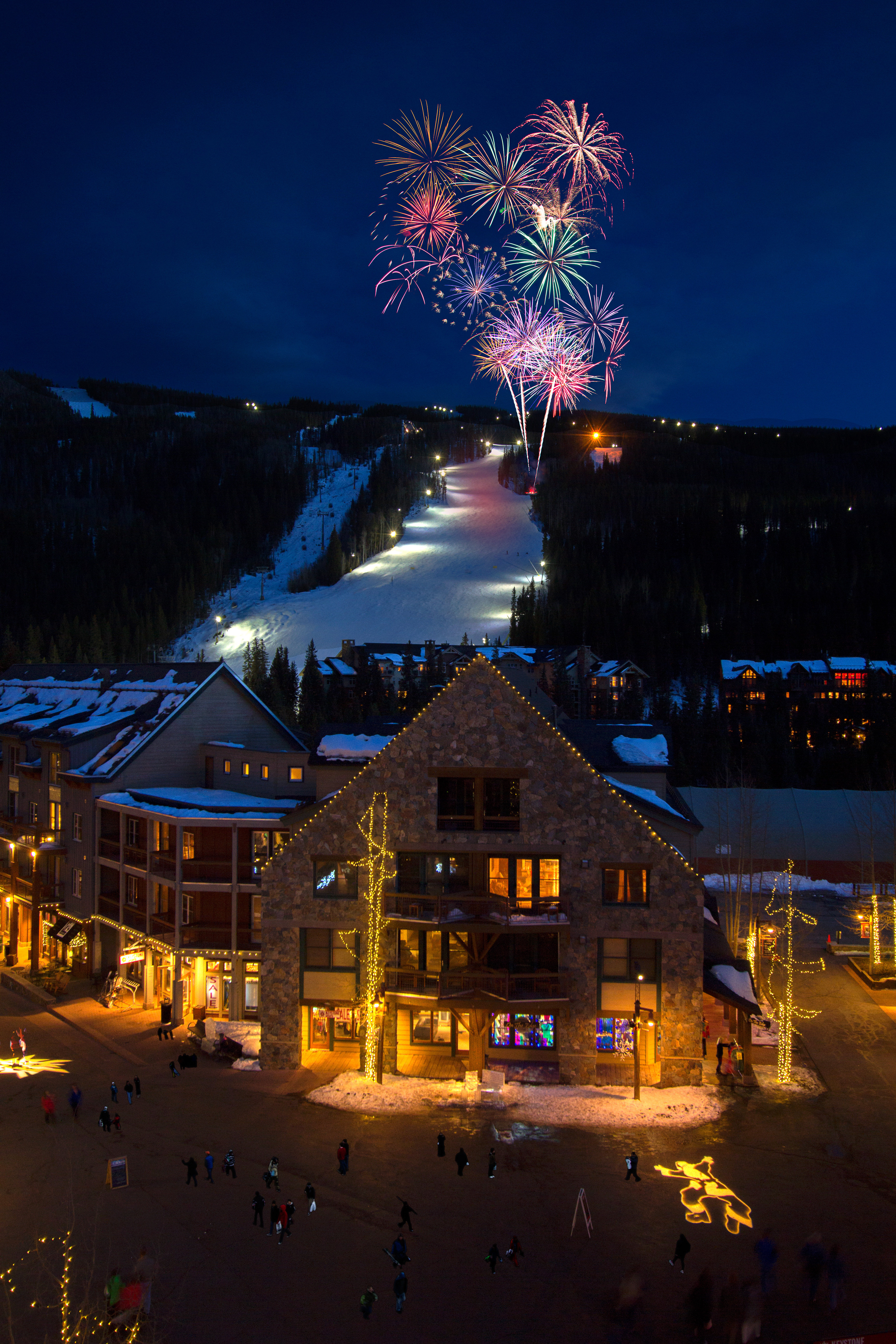 This stunning high-angle exterior view captures a rustic, multi-story lodge in a ski village at night, beautifully illuminated by warm architectural lighting and decorated festive trees. The scene showcases a ski slope in the background with vibrant fireworks blooming in the sky above the mountain. The property presents a quintessential mountain resort aesthetic, perfect for visitors looking for a premium alpine retreat.