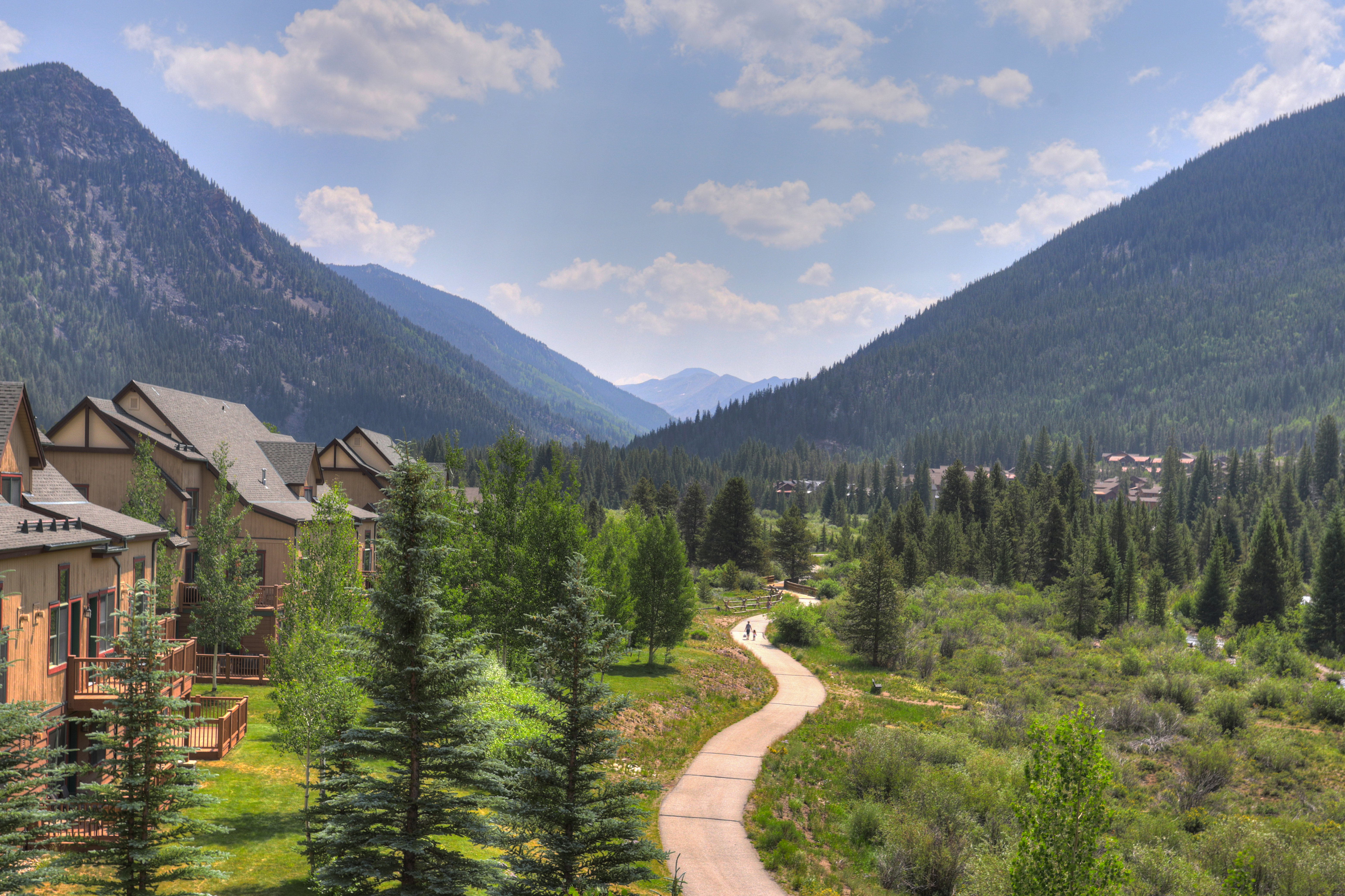 This scenic exterior view captures a mountain resort property, highlighting the multi-family condo buildings with wooden siding and expansive decks. A winding path meanders through the lush, forested valley, surrounded by majestic mountain ridges under a bright, cloudy sky. The setting offers an inviting, serene atmosphere perfect for outdoor enthusiasts seeking proximity to natural trails and mountain landscapes.