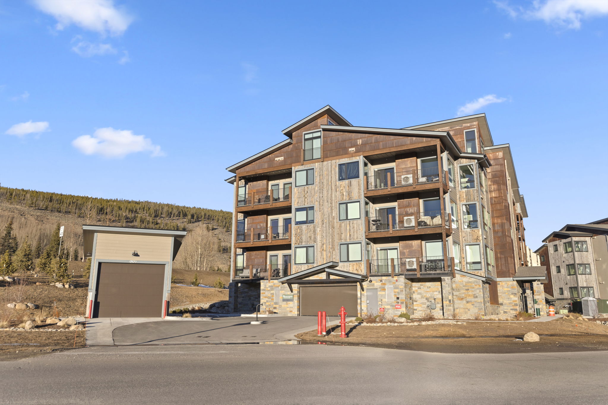 This three-story multi-family residential building showcases a mountain modern architectural style, combining rustic wood siding, stone accents, and metal roofing. A dedicated, detached single-car garage sits gracefully to the side, while balconies provide outdoor living space for residents. The structure overlooks a natural, hilly landscape, projecting a rugged yet upscale appeal perfect for a vacation or mountain environment.
