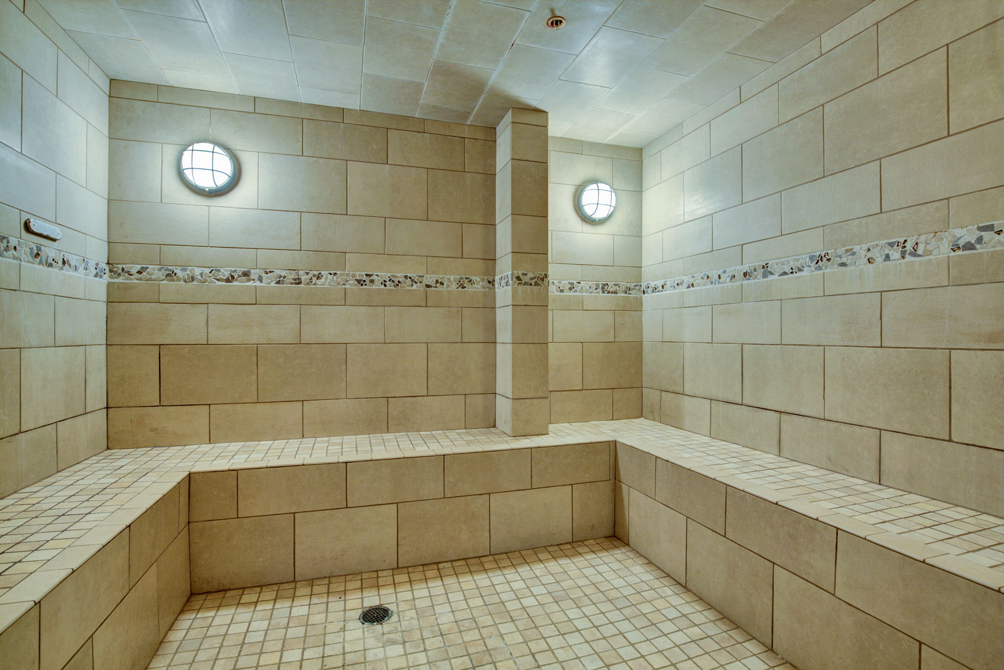 This interior space features a tiled steam room or sauna with L-shaped built-in seating. The room is finished with light beige rectangular wall tiles and square floor tiles, accented by a decorative mosaic tile strip and circular wall-mounted lights. This well-maintained area reflects a high-end amenity typical of luxury apartment complexes or private spa facilities.