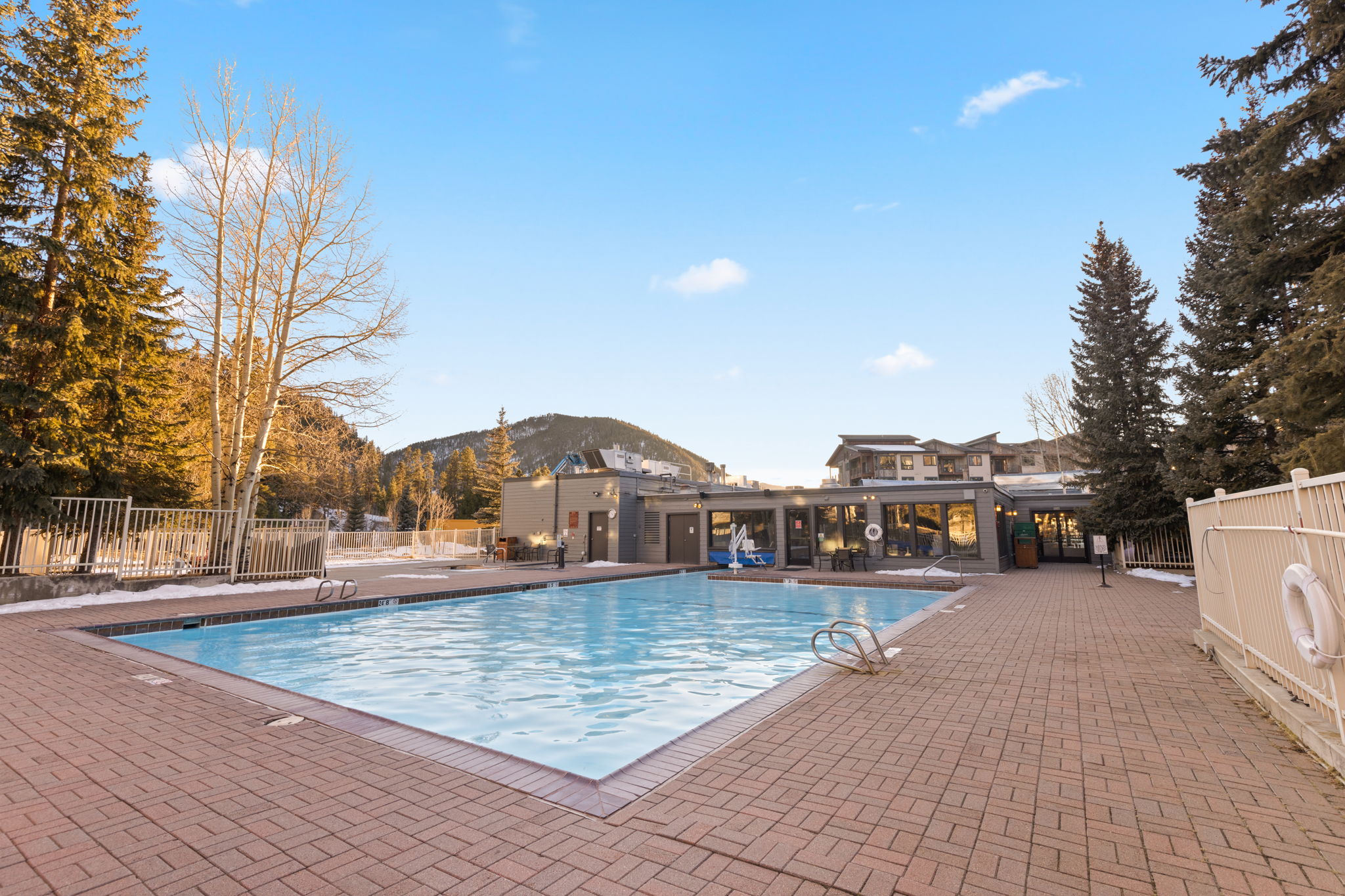 This outdoor swimming pool area features a large, paved pool deck constructed from red bricks, surrounded by a white metal fence and mature pine trees. The pool is positioned directly adjacent to a low-rise building, providing easy access from the property and creating a relaxing communal space. With scenic mountain views in the background and a clear blue sky, the setting offers a clean, inviting atmosphere perfect for leisurely recreation.