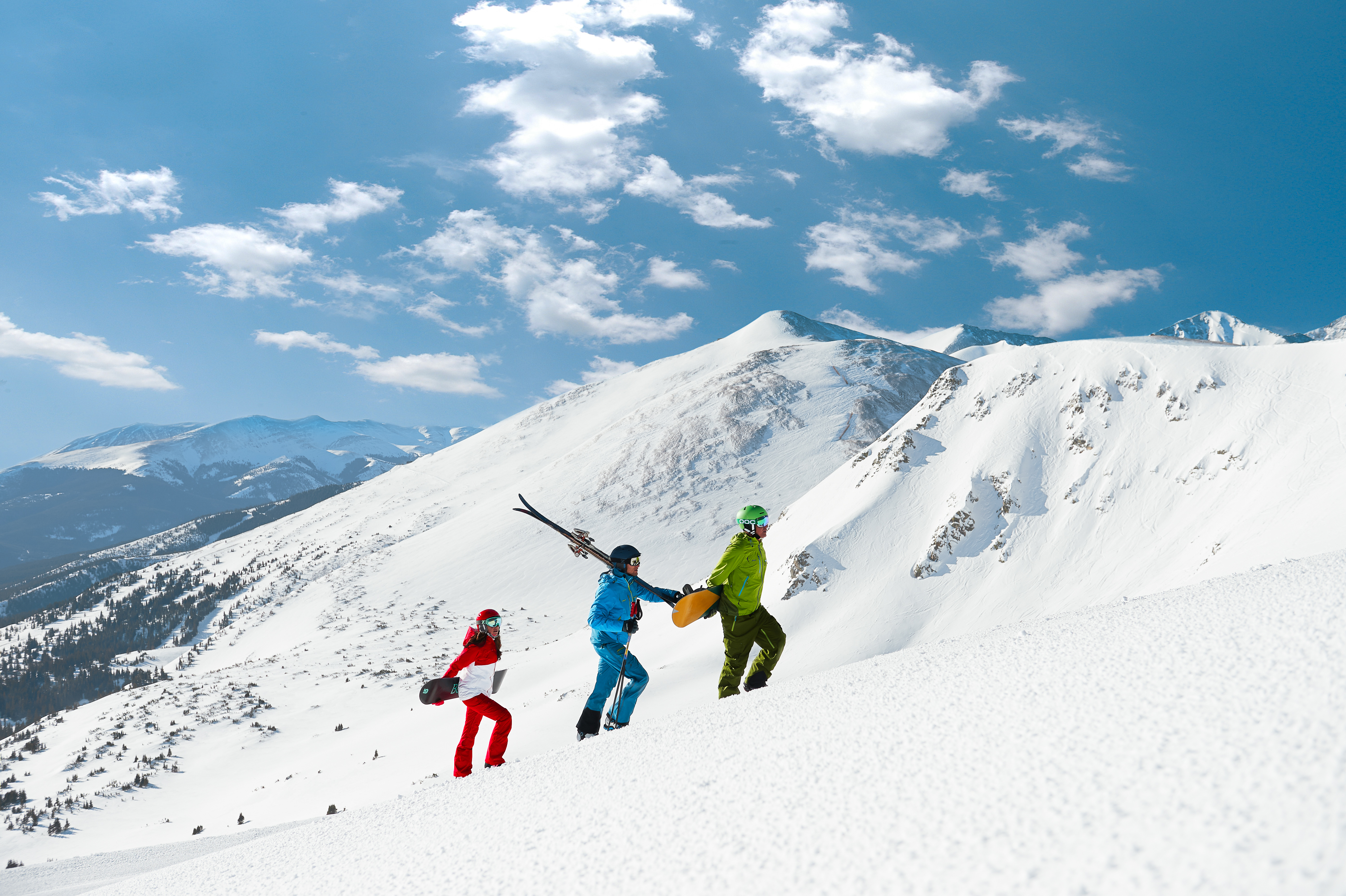 This image shows three individuals hiking up a pristine, snow-covered mountain slope, carrying skiing and snowboarding equipment. The surrounding landscape features vast, rugged alpine terrain under a bright blue sky with scattered clouds, highlighting prime outdoor recreation space. Ideally suited for marketing luxury mountain properties, this shot captures the adventurous lifestyle associated with high-altitude ski-in ski-out real estate.