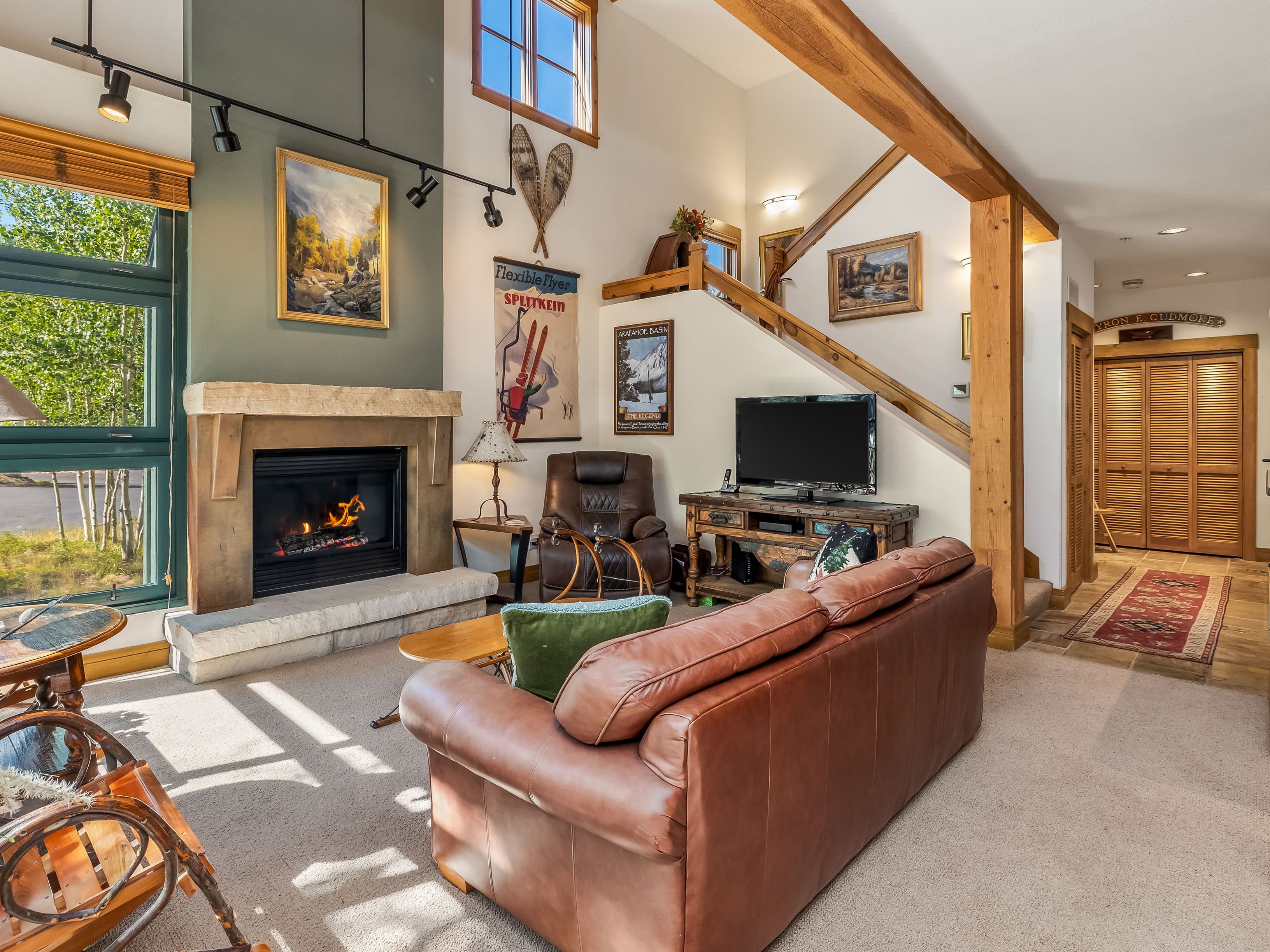 This cozy, rustic-style living room features a prominent stone fireplace with a wood mantel and a tall, sage-colored chimney breast. The space is furnished with a large brown leather sofa, a matching leather armchair, and a wooden media console, all set against a backdrop of vaulted ceilings and exposed timber beams. Large windows to the left provide ample natural light, while vintage-inspired wall art and snowshoes add a charming, mountain-lodge aesthetic to the room.
