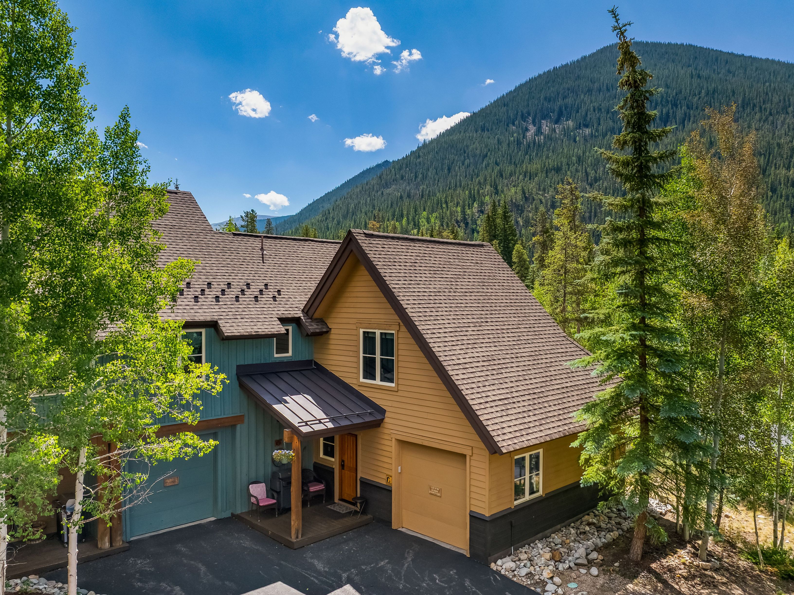 This elevated perspective captures a multi-level mountain home featuring a blend of blue and tan siding with dark brown trim. The property includes two garage bays, a covered entryway, and a dark shingled roof, all set against a backdrop of a lush, forested mountain slope under a clear blue sky. The scene conveys a serene, rustic aesthetic typical of a high-altitude retreat.