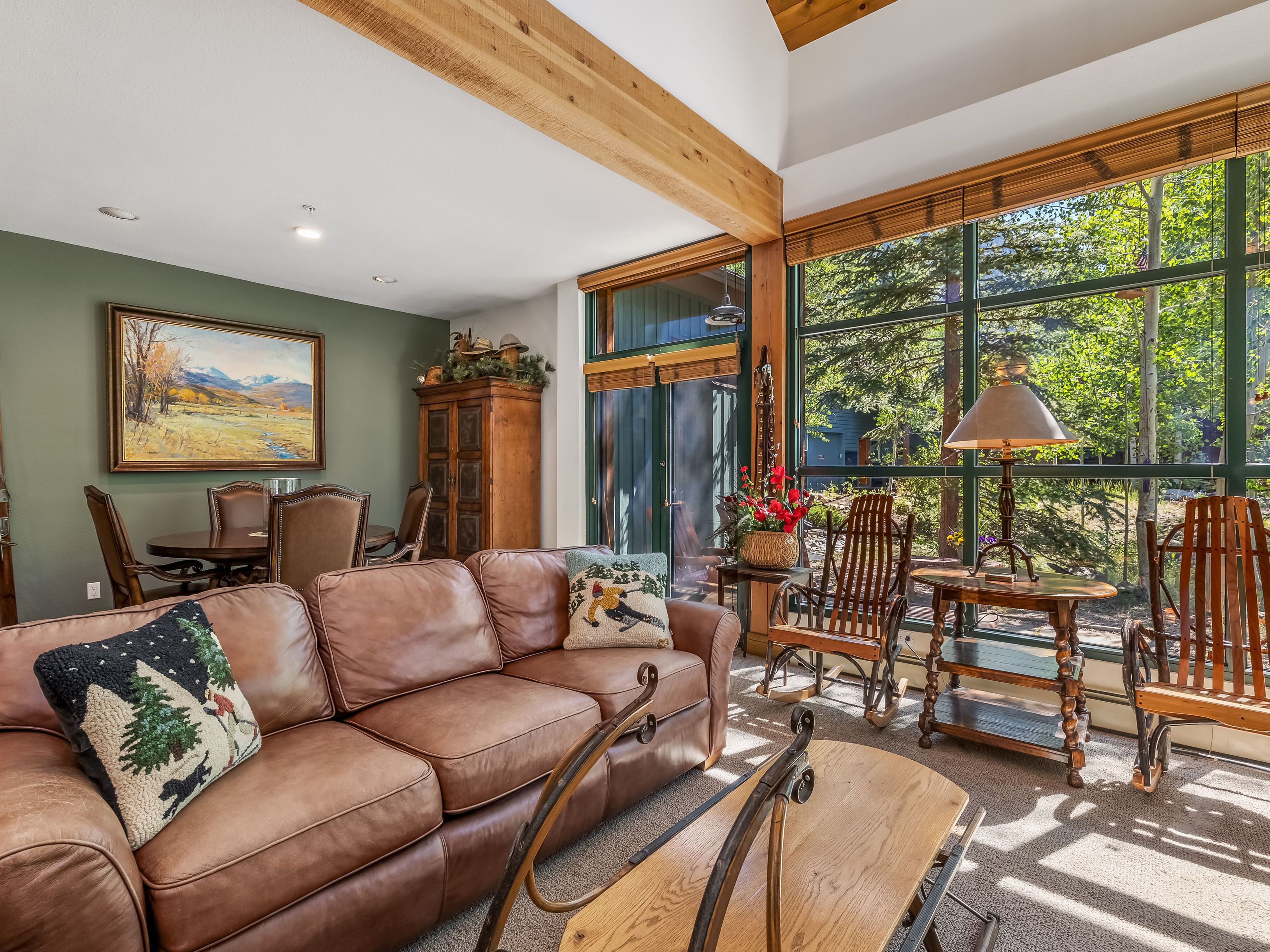This inviting living room features a large brown leather sofa adorned with decorative pillows, positioned in front of a unique wooden coffee table. The space is bathed in natural light from expansive windows that offer a view of the surrounding trees, and it includes a cozy seating area with two wooden rocking chairs and a side table with a lamp. A dining area with a wooden table and chairs is visible in the background, set against a green accent wall with a landscape painting, creating a warm and rustic atmosphere.