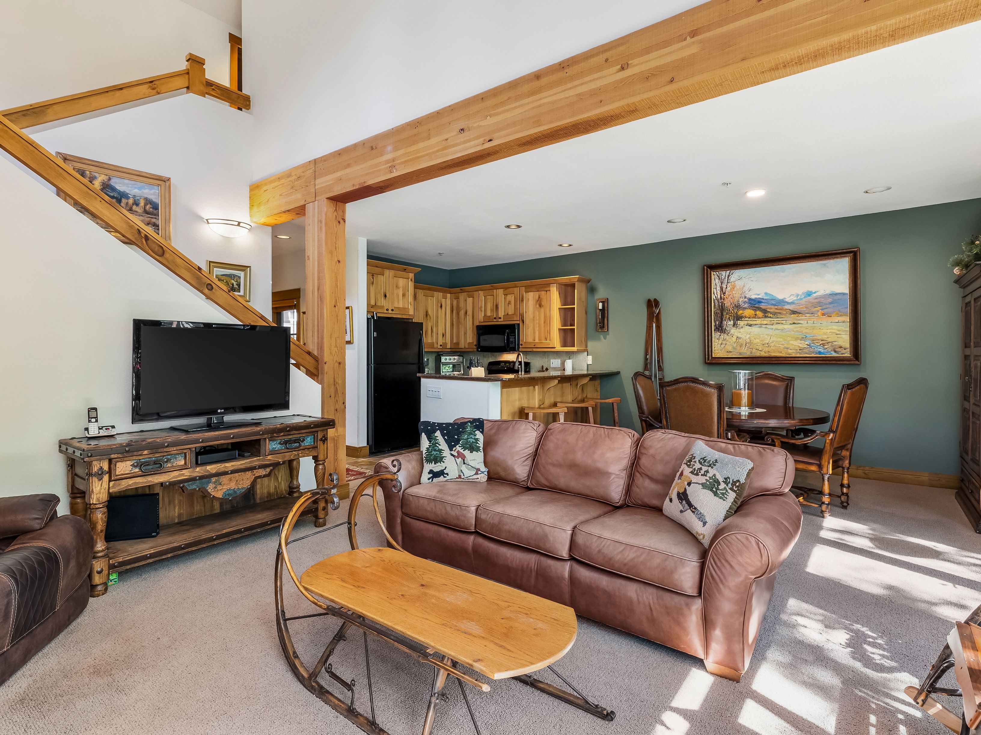 This open-concept living area features a rustic mountain aesthetic with a prominent exposed wooden beam structure and a staircase leading to an upper level. The space includes a brown leather sofa, a unique coffee table fashioned from a vintage wooden sled, and a media console, all set against a backdrop of sage green walls and a dining area. The kitchen is visible in the background, showcasing light wood cabinetry and a breakfast bar, creating a cohesive and inviting living space.