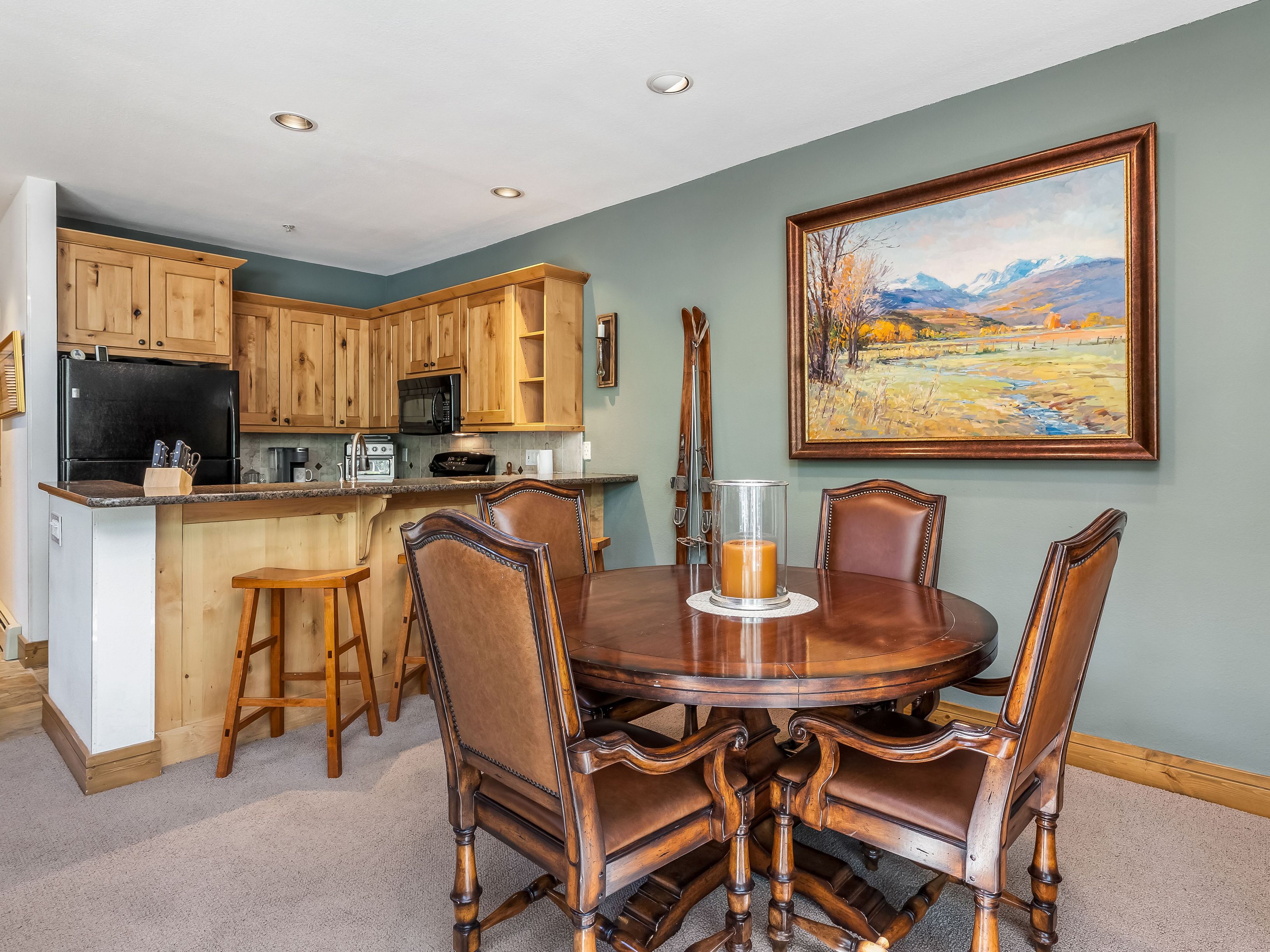 This open-concept dining area features a round wooden table surrounded by four leather-upholstered chairs, positioned adjacent to a kitchen breakfast bar. The space is decorated with a large landscape painting on the wall and a pair of vintage wooden skis, creating a cozy, mountain-inspired aesthetic. The kitchen area includes light wood cabinetry and a black refrigerator, providing a functional and warm transition between the cooking and dining spaces.