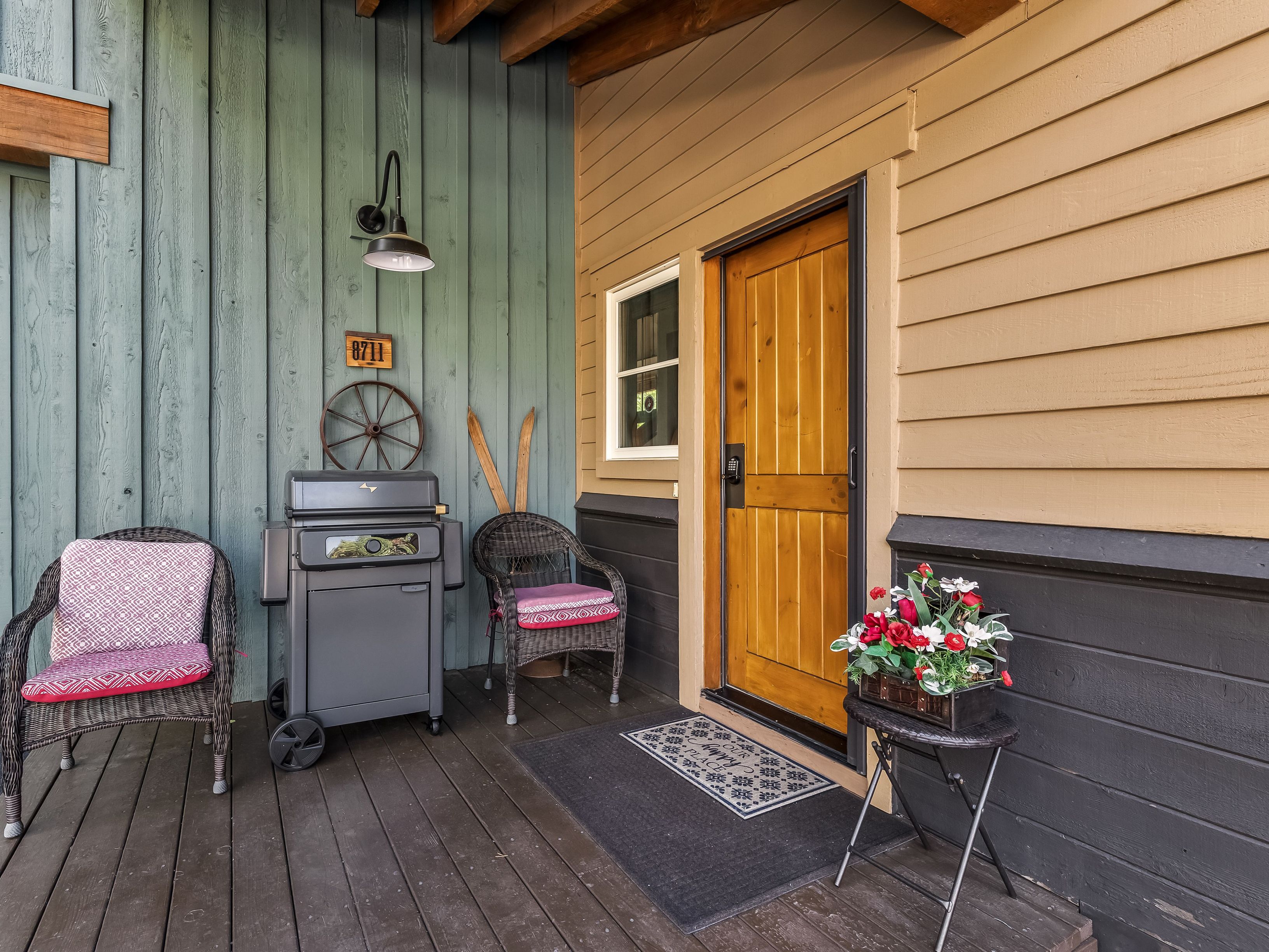 This inviting entryway features a rustic wooden door set against a mix of teal vertical siding and tan horizontal lap siding. The covered porch area includes two wicker chairs with patterned cushions, a charcoal-colored grill, and a small side table holding a vibrant floral arrangement. A welcome mat sits before the door, which is accented by a wall-mounted light fixture and decorative vintage-style wall decor.