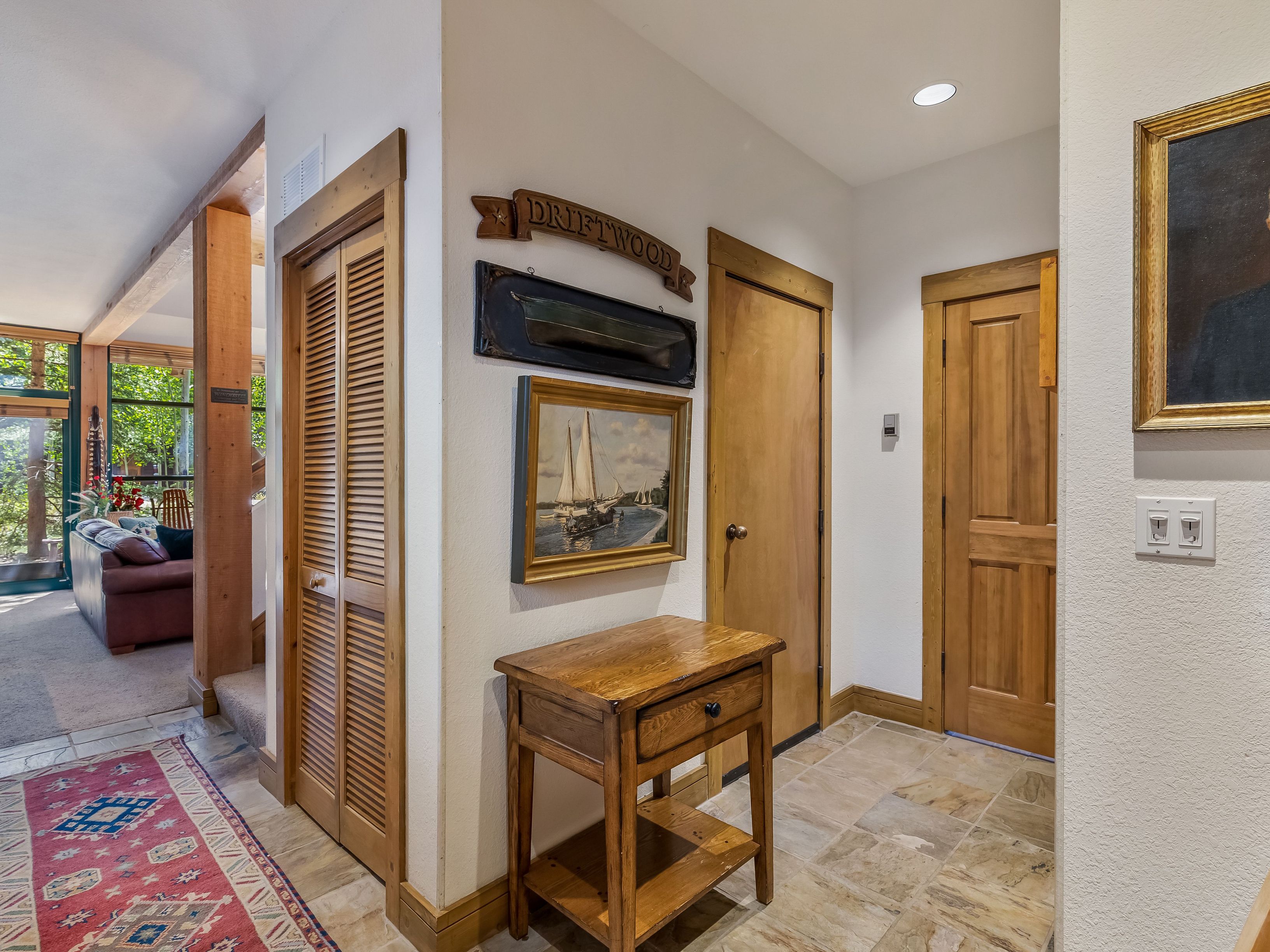 This interior hallway features light-colored walls, stone tile flooring, and warm wood trim throughout. The space includes a small wooden side table, a decorative 'DRIFTWOOD' sign, and framed nautical artwork, leading toward a glimpse of a living area with a sofa and large windows. The perspective is from the hallway, looking past the wooden doors and decor into the adjacent room.