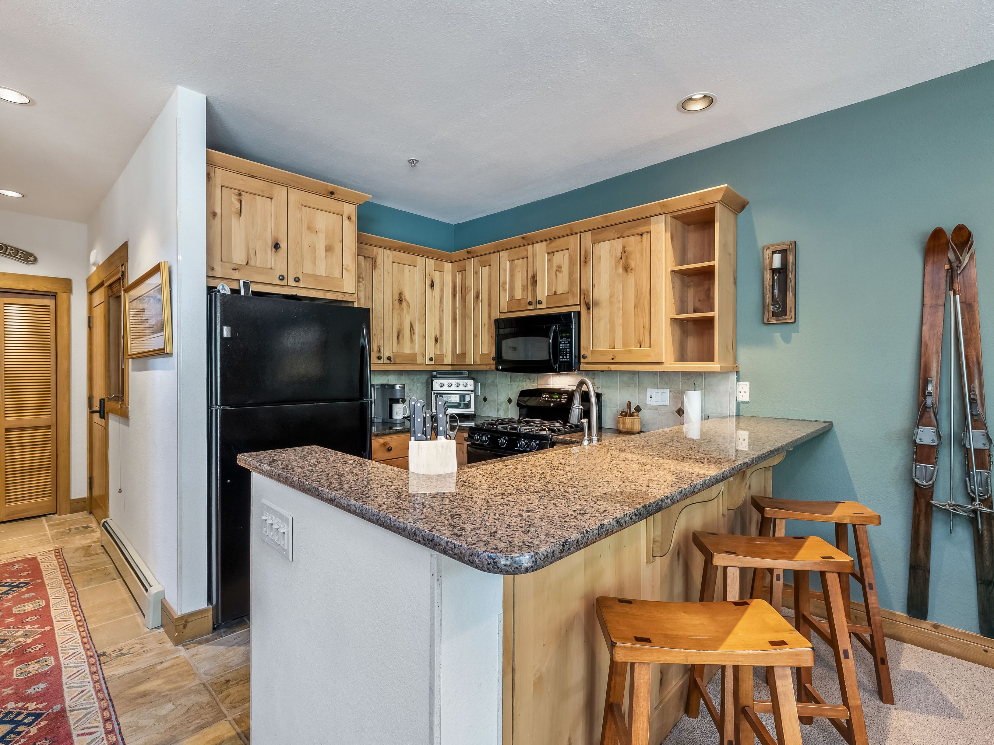 This cozy kitchen features light wood cabinetry, a dark granite countertop, and black appliances, creating a rustic mountain aesthetic. A breakfast bar with three wooden stools provides casual seating, while vintage wooden skis mounted on the teal-painted wall add a unique decorative touch. The perspective captures the functional layout of the kitchen, highlighting the transition from the tiled entryway to the carpeted living area.