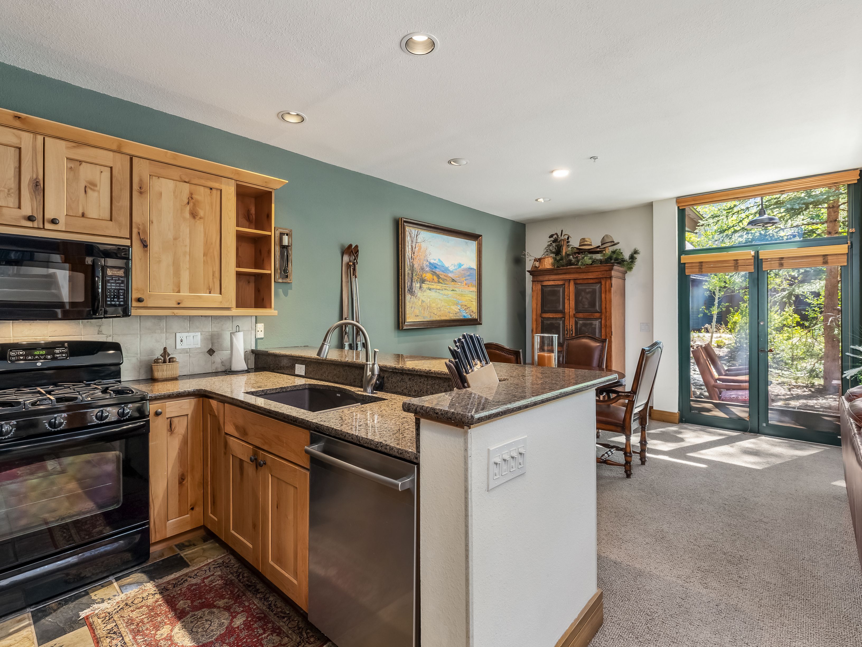 This kitchen features light wood cabinetry, granite countertops, and stainless steel appliances, including a microwave and dishwasher. The space is open to a dining area with a wooden hutch and a view through large glass doors that lead to a wooded outdoor area. The room is accented by a green wall, a patterned rug, and recessed lighting, creating a warm and inviting atmosphere.