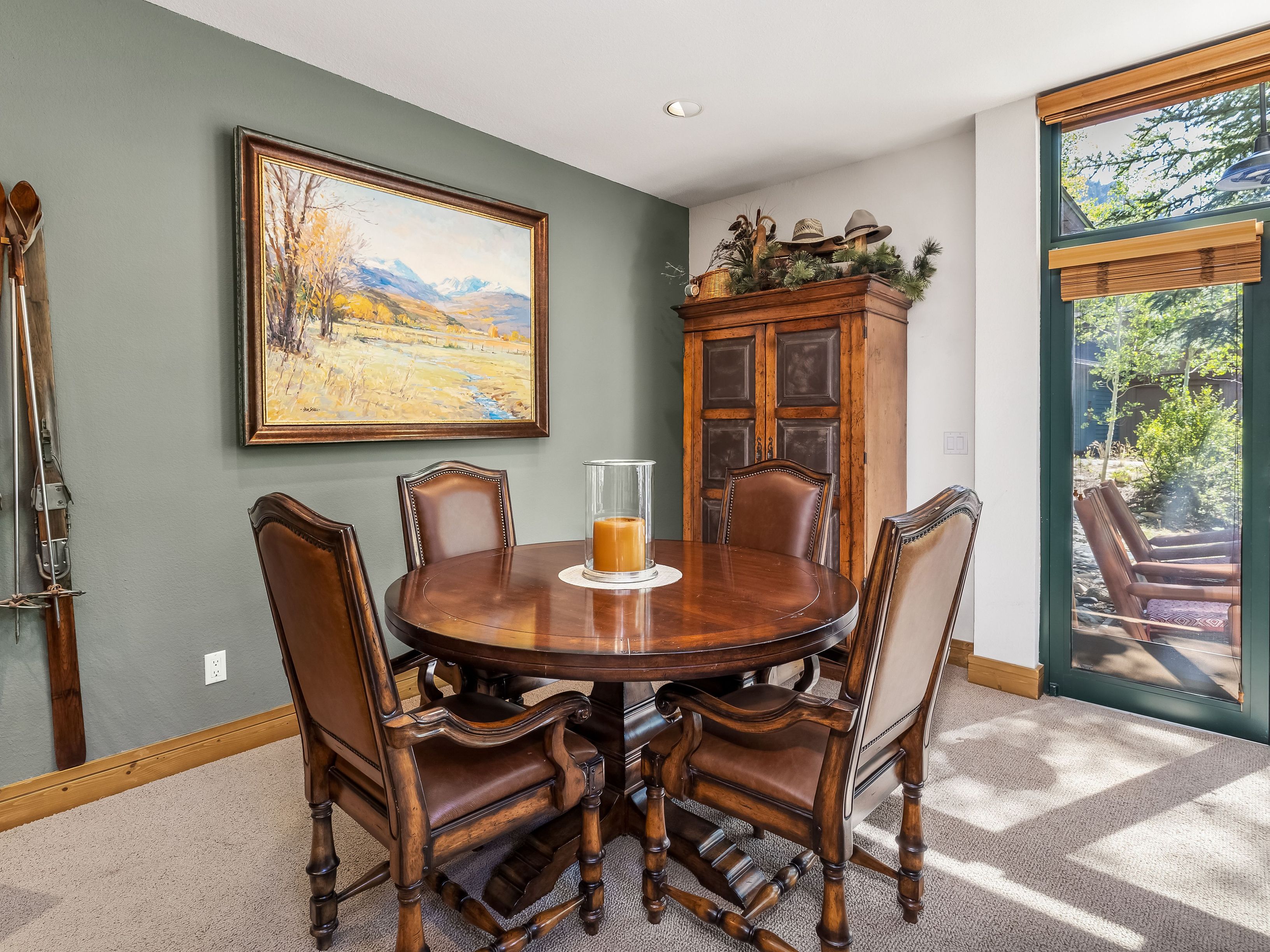 This dining area features a round, dark wood table surrounded by four matching leather-upholstered chairs, creating a warm and traditional atmosphere. A large landscape painting hangs on the sage green wall, while a tall wooden cabinet provides storage and decorative display space. The room is illuminated by natural light streaming through a large window that offers a view of the outdoor greenery.