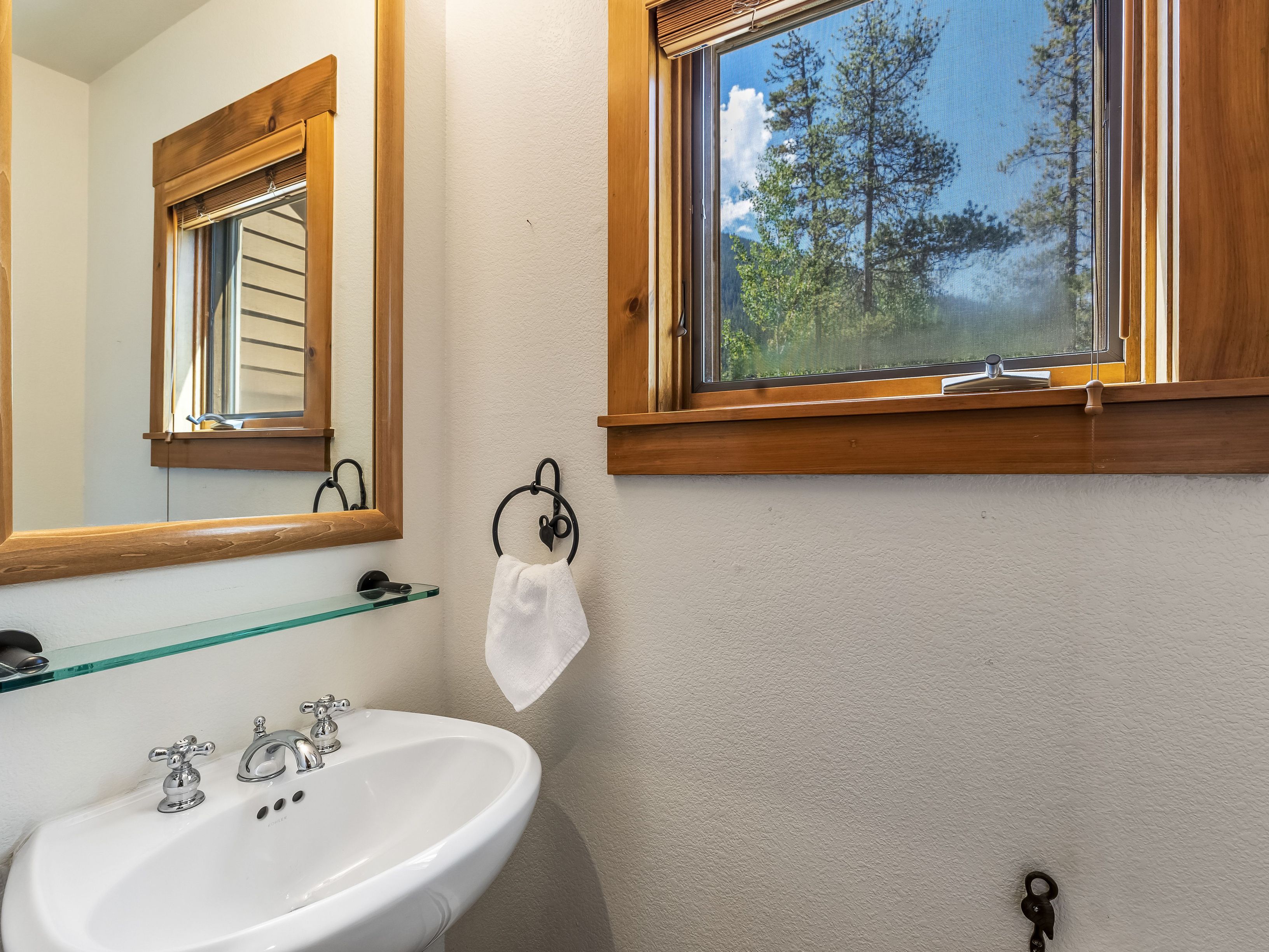 This compact powder room features a white pedestal sink with polished chrome fixtures and a glass shelf mounted above. A large mirror with a warm wood frame reflects the window, while a matching wood-framed window provides a view of the outdoors. The space is finished with neutral walls and a dark metal towel ring, creating a clean and rustic aesthetic.