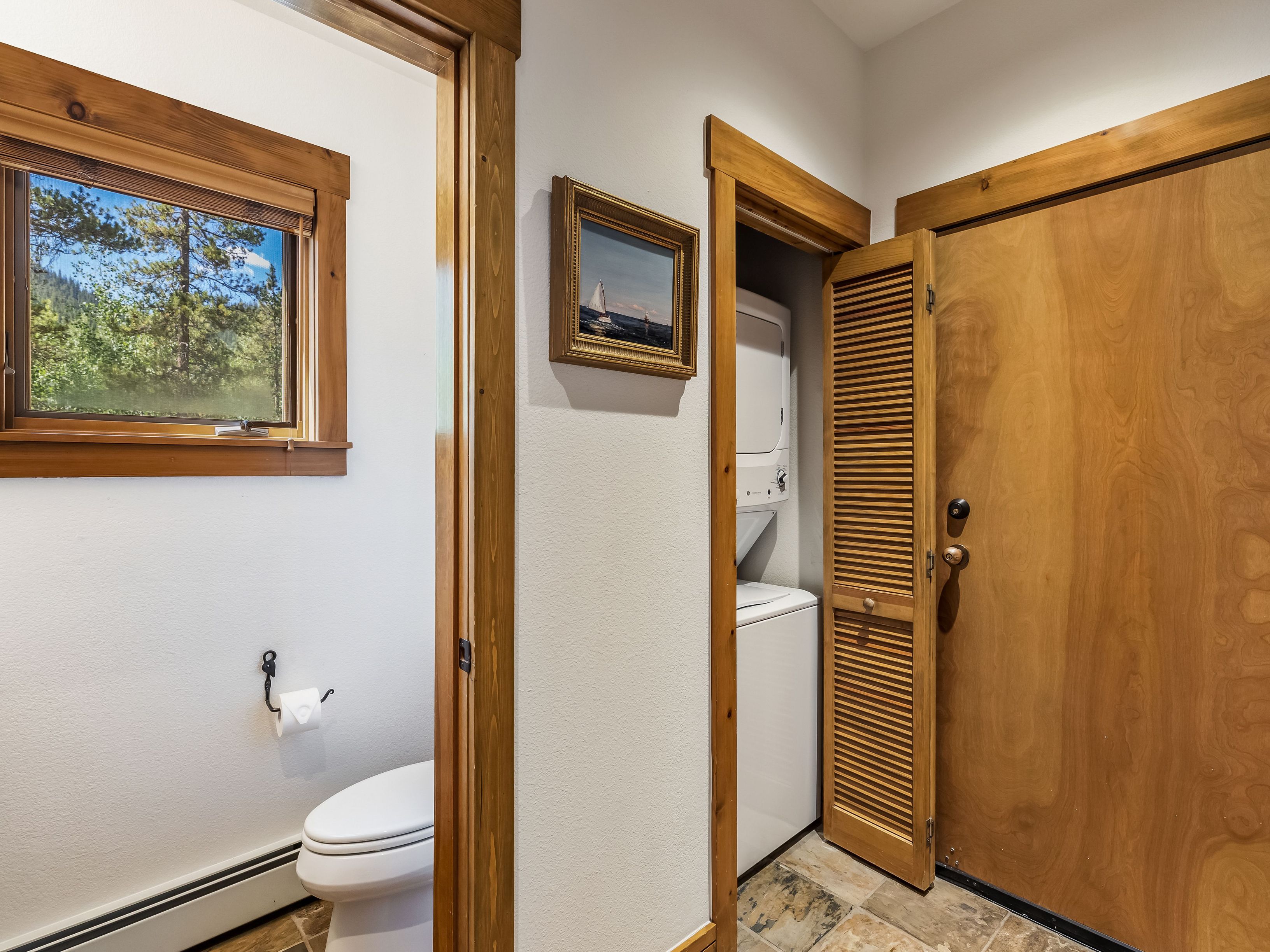This interior view captures a functional laundry area featuring a stacked washer and dryer unit tucked behind a louvered wooden door. To the left, a doorway leads into a bathroom equipped with a toilet and a window that provides a view of the surrounding trees. The space is finished with light-toned walls, warm wood trim, and patterned tile flooring, creating a rustic and practical atmosphere.