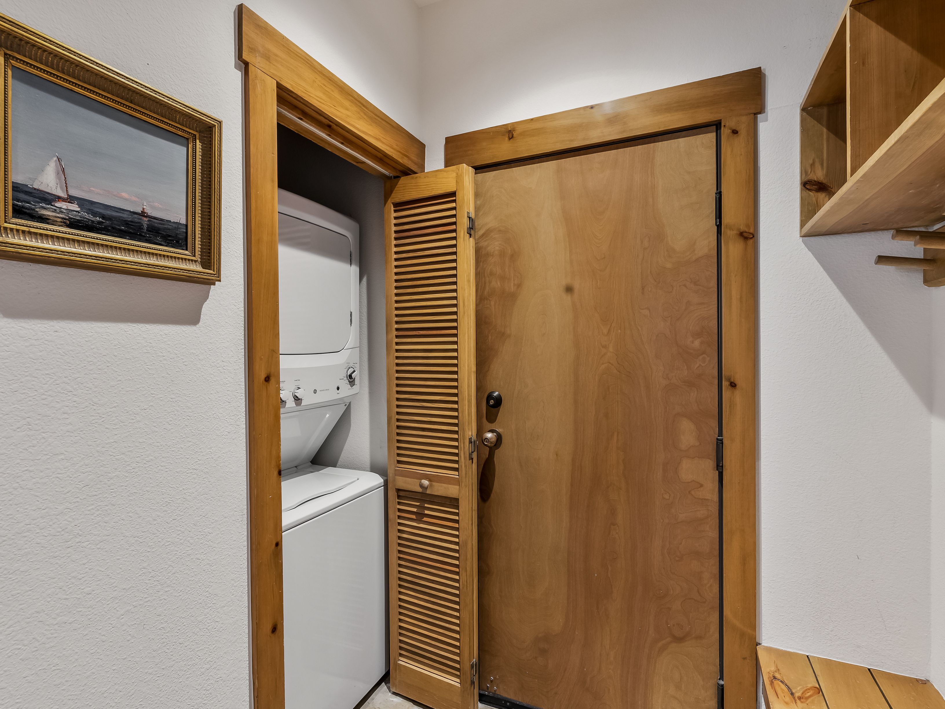 This interior view shows a compact laundry area featuring a stacked washer and dryer unit tucked into a closet space. The area is framed by light wood trim and includes a louvered door, a wooden door leading to another room, and a wall-mounted wooden shelf. A framed nautical painting hangs on the wall to the left, adding a decorative touch to the functional space.