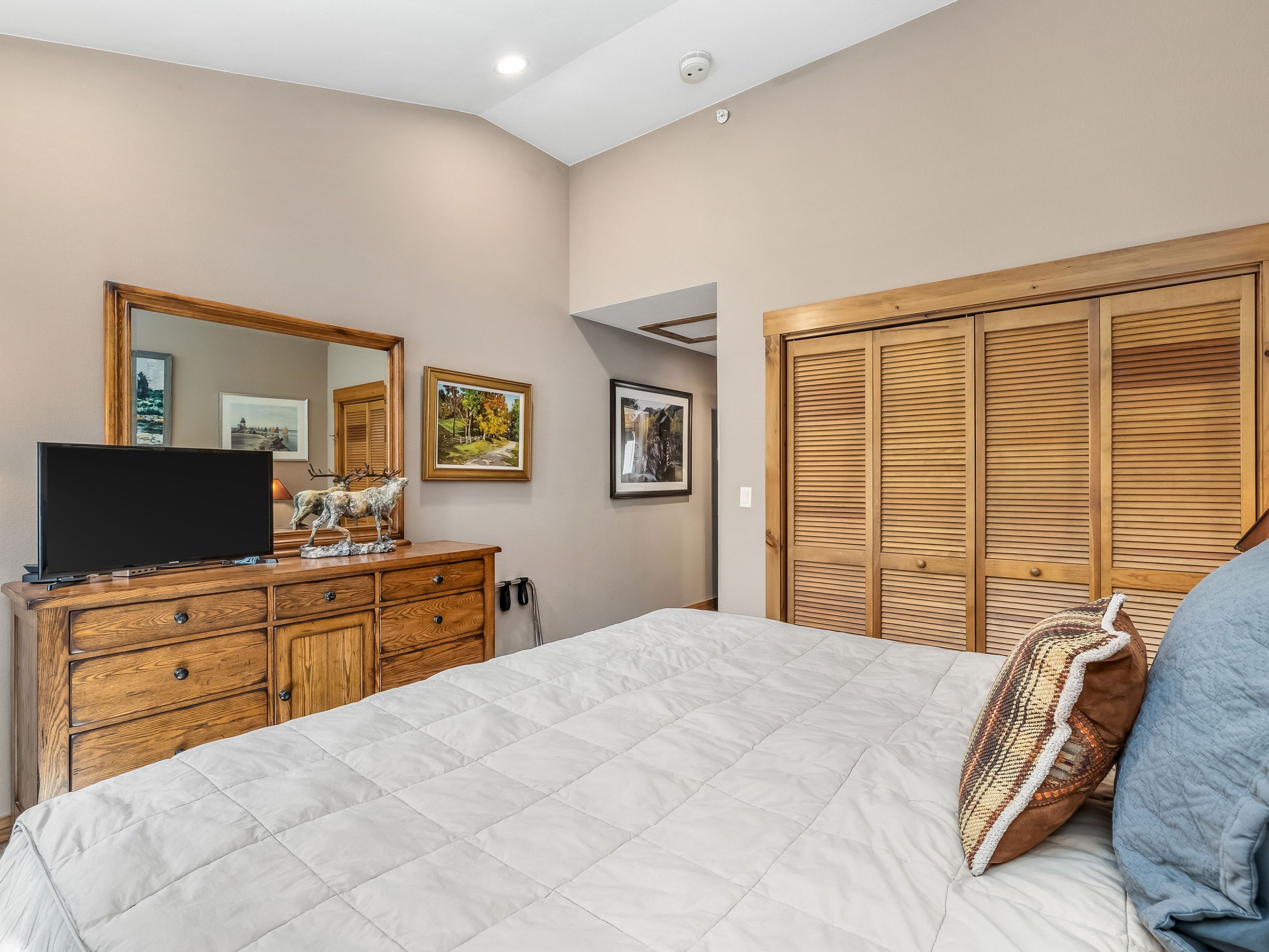 This bedroom features a neutral color palette with a large wooden dresser topped by a mirror and decorative animal figurines. A bed with a light-colored quilted comforter and patterned accent pillows occupies the foreground, while louvered wooden closet doors provide ample storage space. The room is accented with framed artwork on the walls and has a vaulted ceiling, creating a cozy and traditional atmosphere.