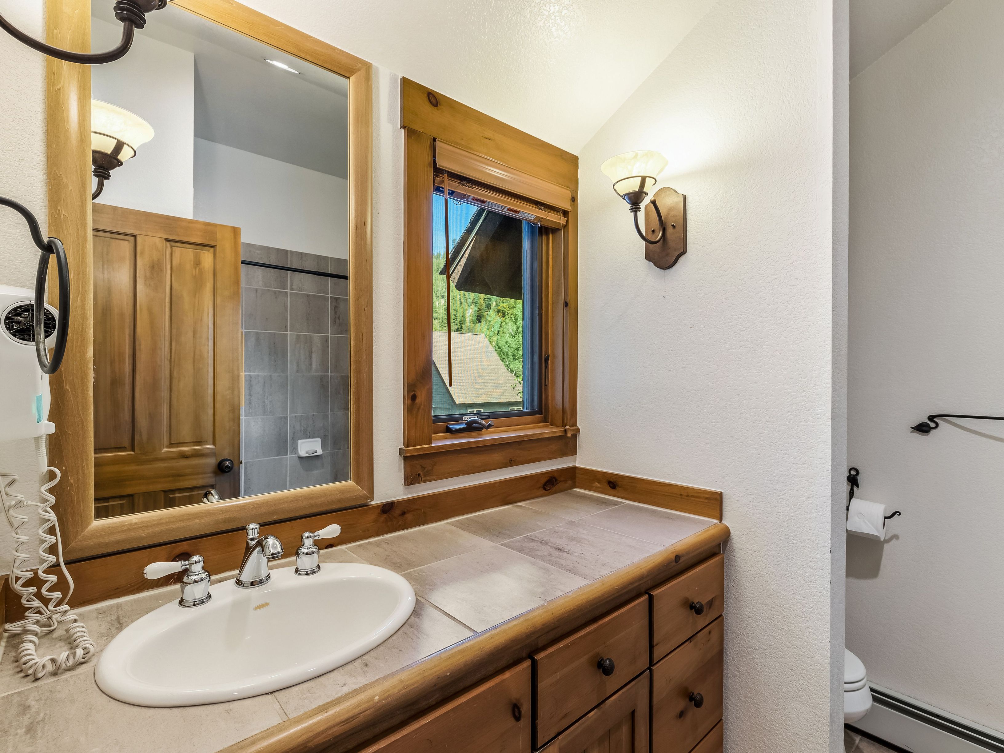 This rustic-style bathroom features a wooden vanity with a tiled countertop and an oval drop-in sink. A large mirror with a matching wood frame hangs above the vanity, reflecting a tiled shower area, while a window with wooden trim provides natural light. The space is completed by a wall-mounted light fixture and a separate toilet area visible to the right.