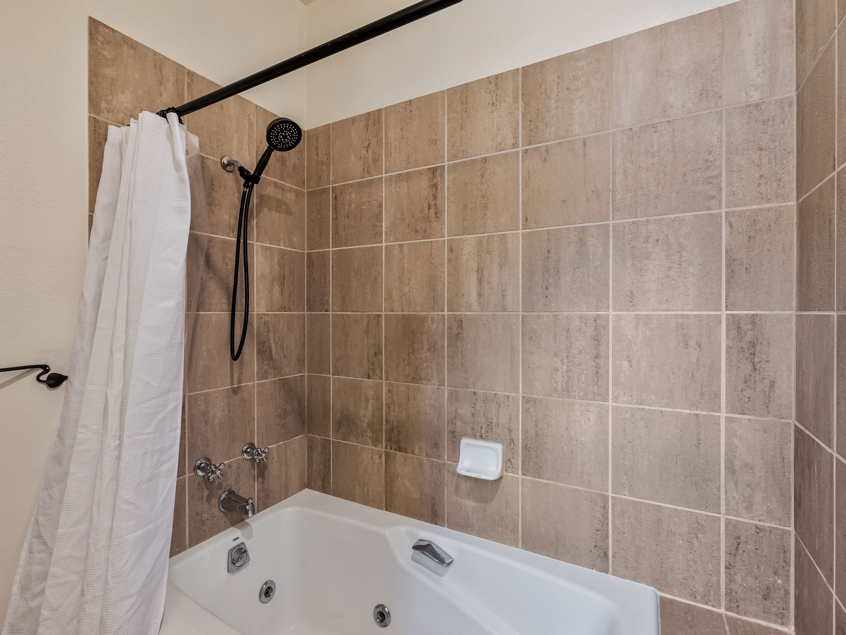 This bathroom features a jetted soaking tub set against a wall tiled with large, neutral-toned square tiles. A black shower fixture with a handheld sprayer is mounted on the wall, complemented by a matching black curtain rod and a white shower curtain. A small, white soap dish is mounted on the tiled wall, providing a functional touch to the bathing area.