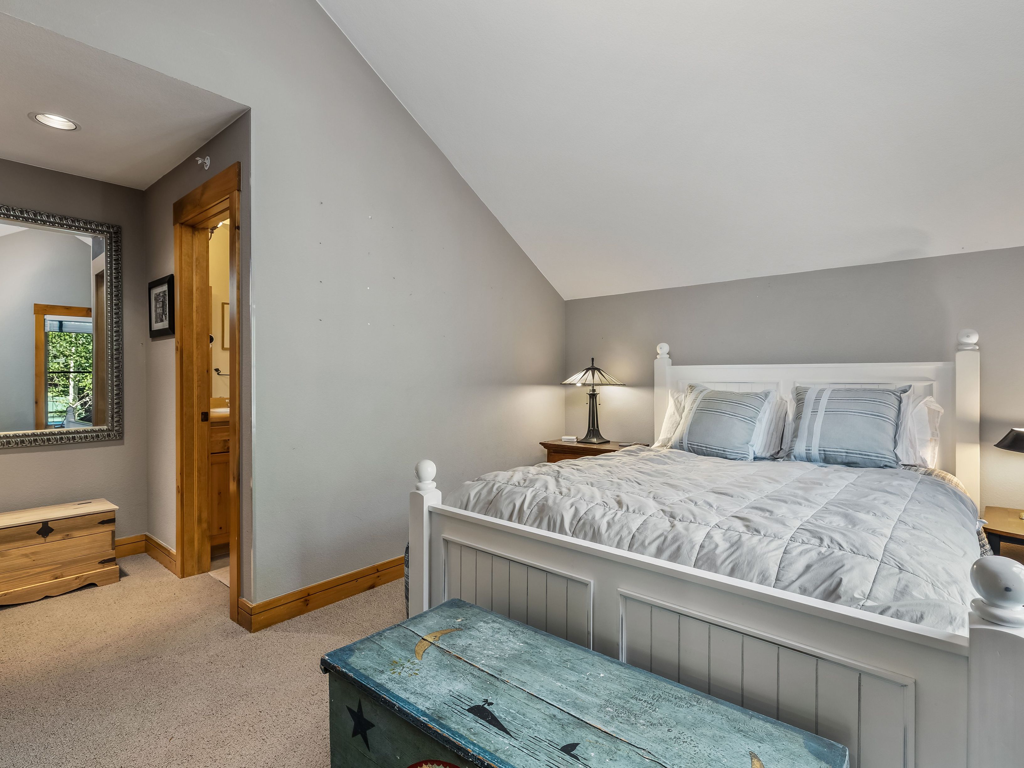 This cozy bedroom features a white wooden bed frame dressed in a light blue quilted comforter and matching pillows. A painted wooden chest sits at the foot of the bed, while a small nightstand with a lamp provides warm lighting against the soft gray walls. To the left, a doorway leads into an adjacent bathroom, and a large mirror reflects the room's interior, adding depth to the space.