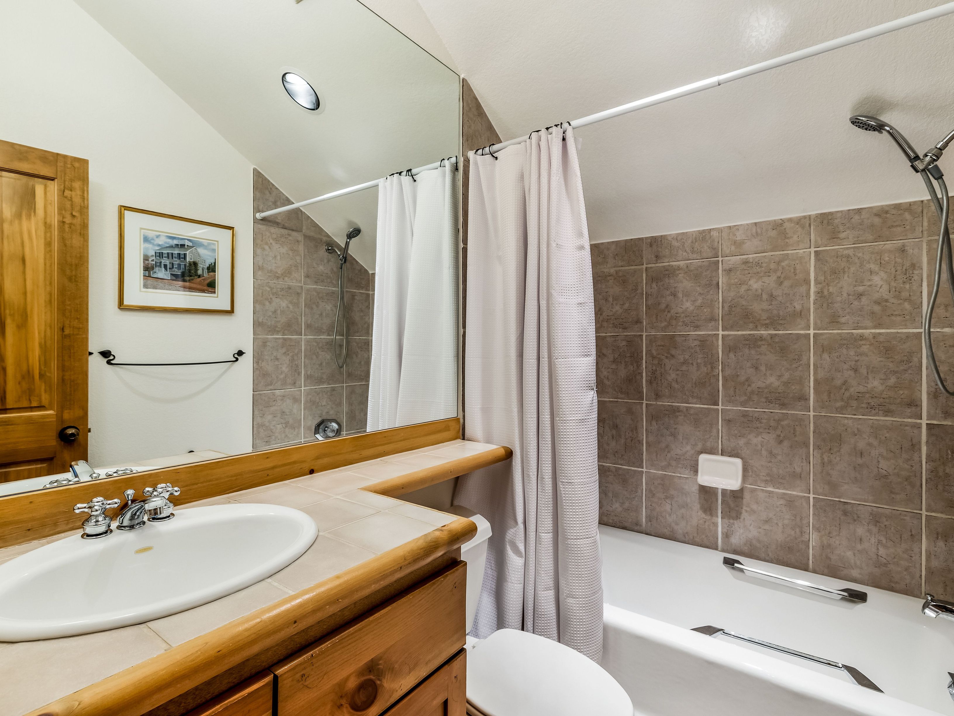 This bathroom features a rustic aesthetic with a light wood vanity topped with neutral-toned tiles and a white drop-in sink. A large mirror spans the wall above the vanity, reflecting the shower area which is enclosed by a white curtain and finished with square, earth-toned wall tiles. The space includes a white toilet and a shower head mounted on the tiled wall, creating a functional and compact layout.