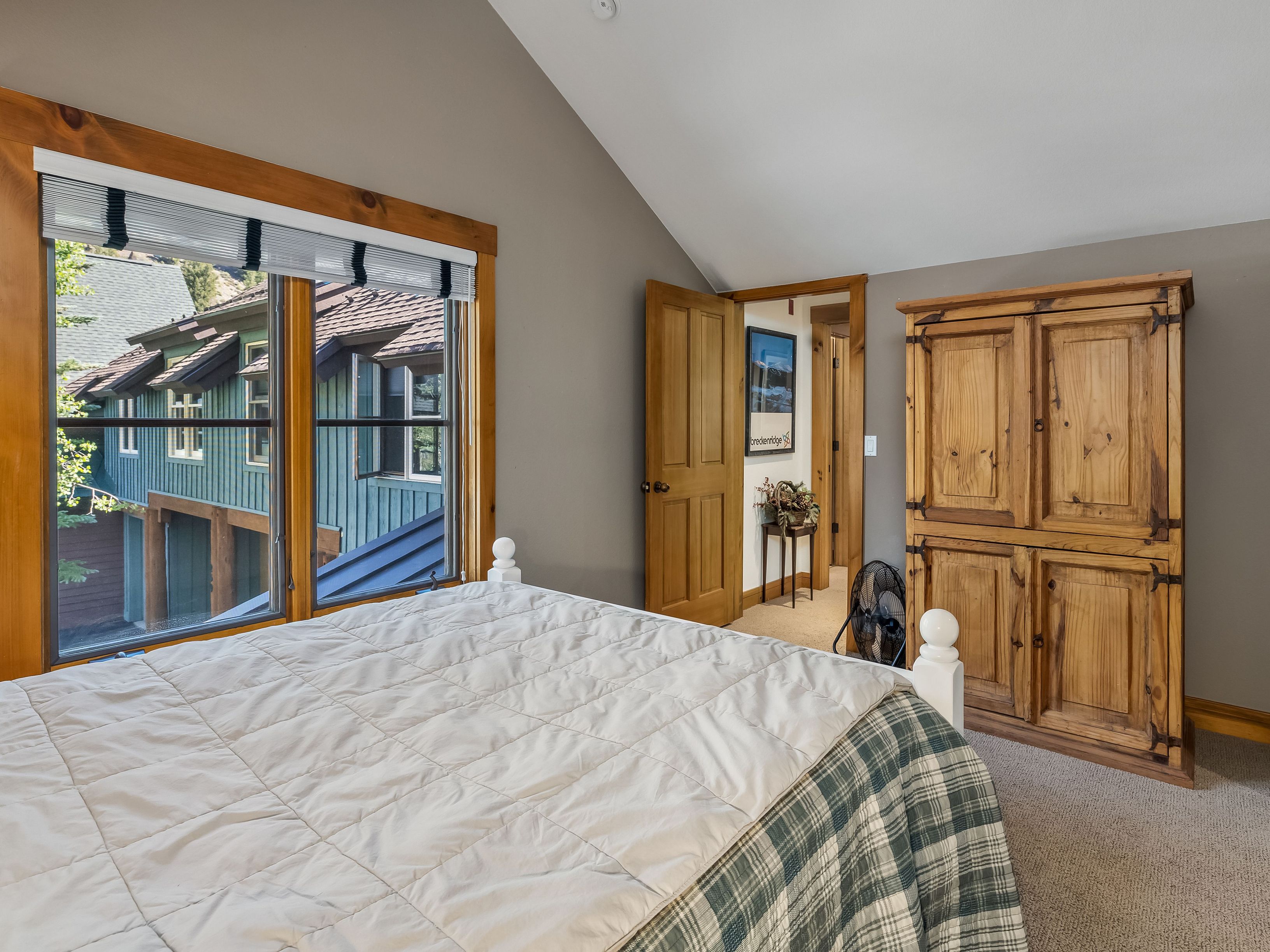 This bedroom features a cozy, rustic aesthetic with a white-framed bed topped with a quilted white coverlet and a green plaid blanket. A large wooden armoire provides storage, while a window with a Roman shade offers a view of the neighboring building's exterior. The room has a vaulted ceiling and neutral wall tones, creating a comfortable and inviting atmosphere.