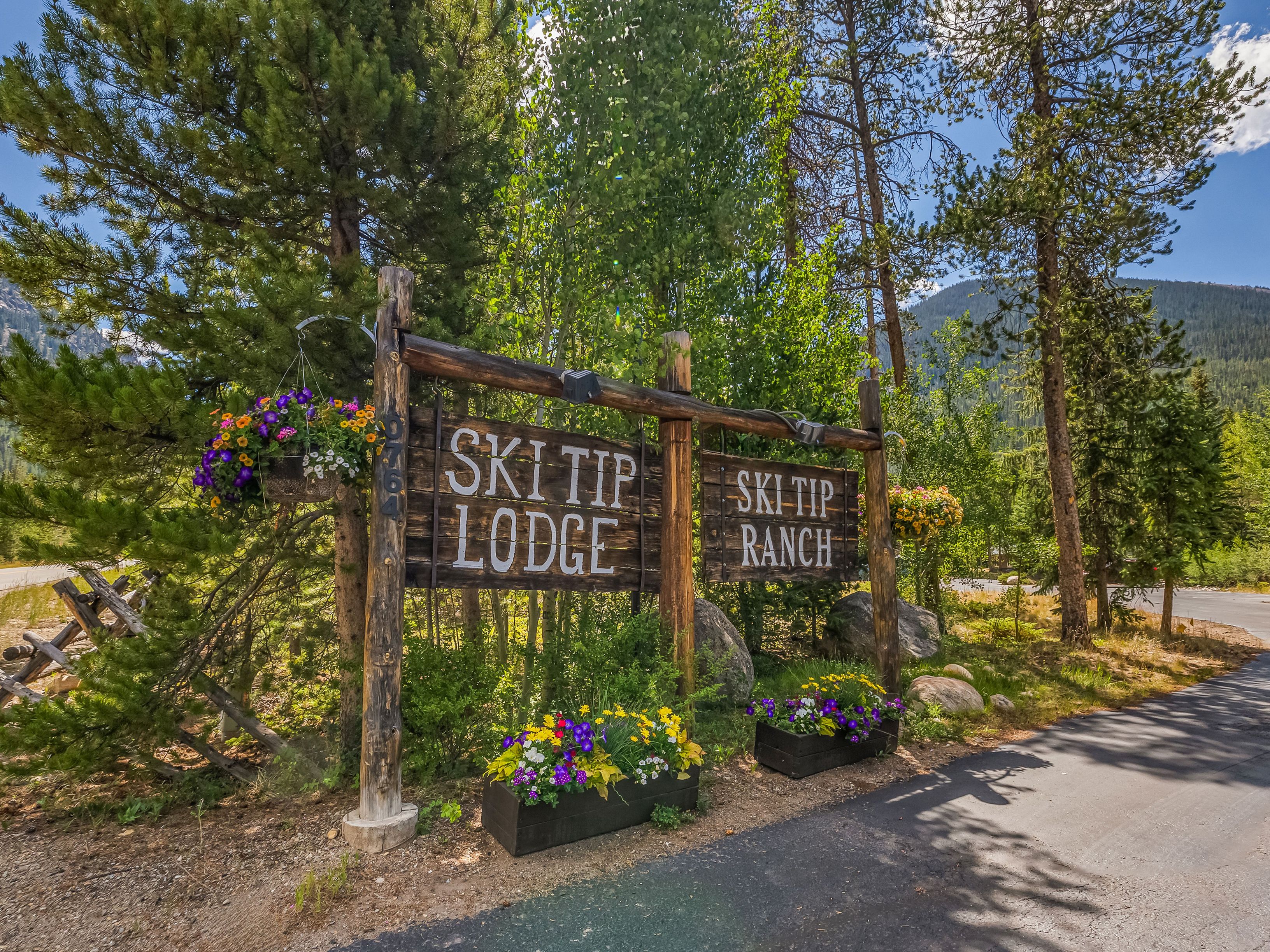 This image features a rustic, wooden signpost marking the entrance to 'Ski Tip Lodge' and 'Ski Tip Ranch,' set against a backdrop of lush pine trees and a clear blue sky. The sign is adorned with vibrant hanging flower baskets and matching planter boxes at its base, creating a welcoming and picturesque arrival point. The perspective is a low-angle, eye-level shot that emphasizes the natural, mountainous setting of the property.