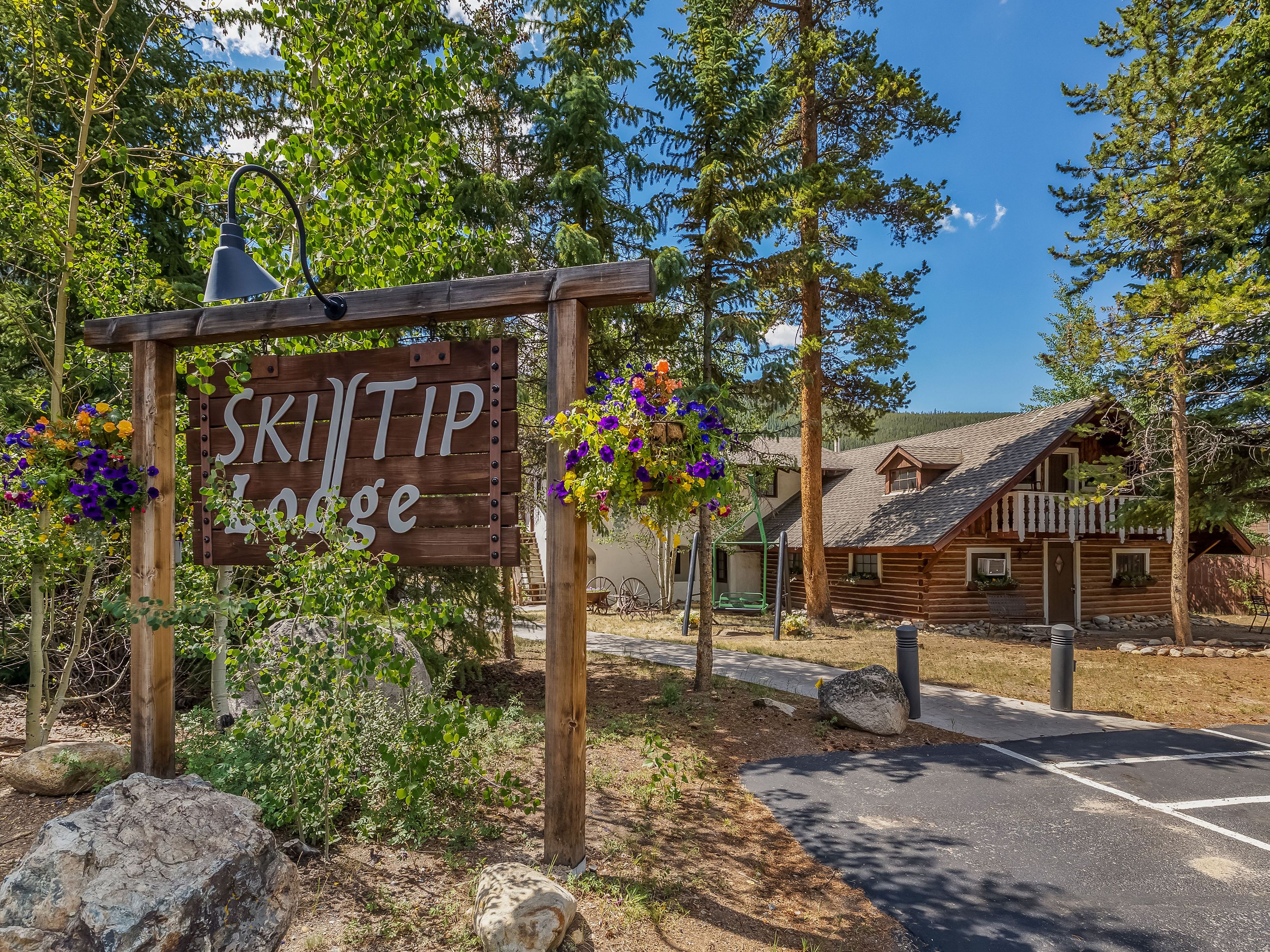 This exterior view features a rustic log-style lodge nestled among tall pine and aspen trees under a bright blue sky. A prominent wooden sign in the foreground identifies the property as 'Ski Tip Lodge,' accented by hanging flower baskets, while a paved path leads toward the main building. The lodge itself showcases traditional log construction, a gabled roofline, and a second-story balcony, creating a welcoming and classic mountain retreat atmosphere.