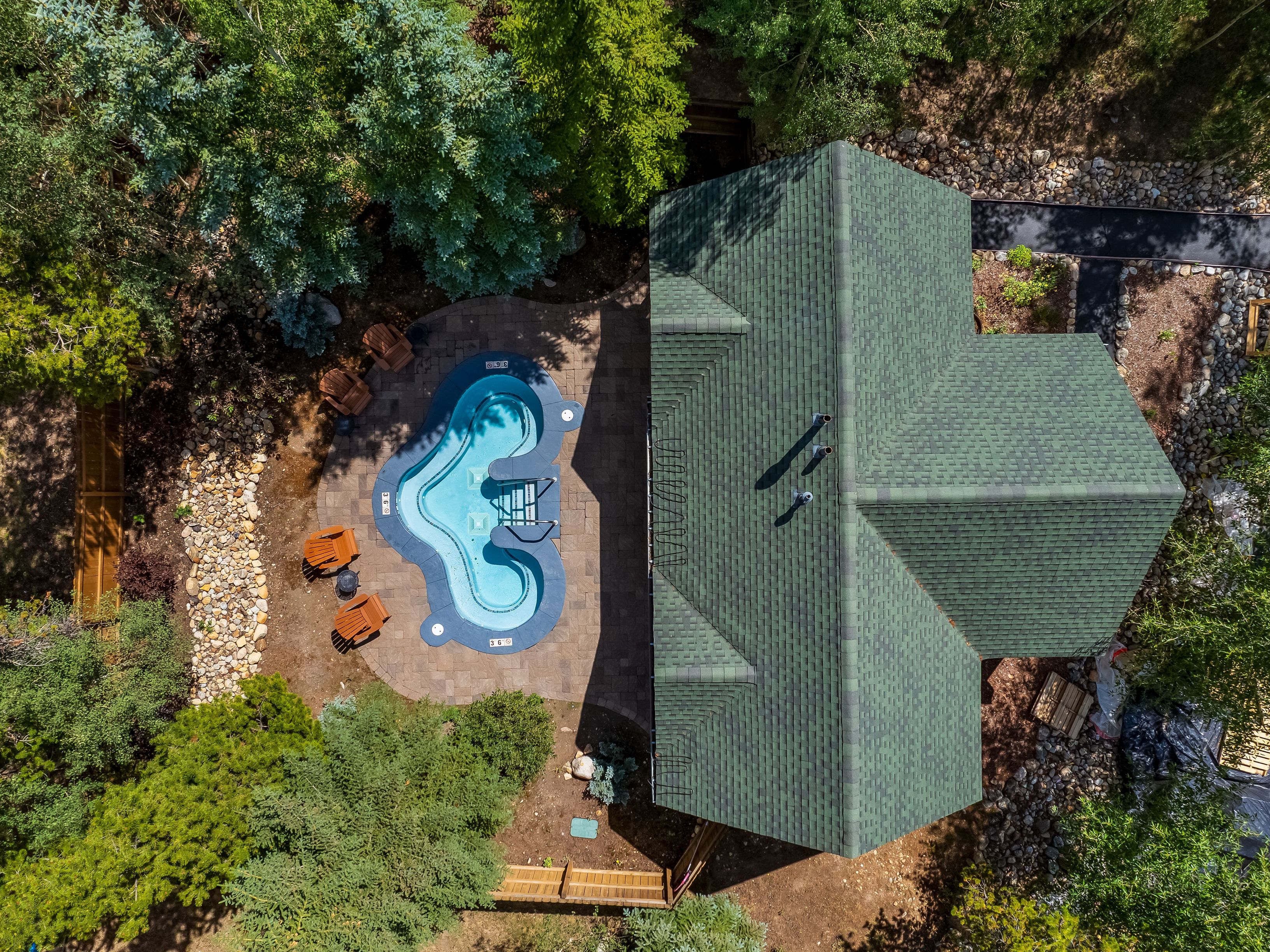 This high-angle aerial perspective captures a property nestled within a lush, wooded landscape. The scene features a uniquely shaped, blue-tiled swimming pool set into a stone-paved patio, surrounded by several wooden lounge chairs. The adjacent building is topped with a dark green shingled roof, emphasizing the home's integration into its natural, forest-like environment.