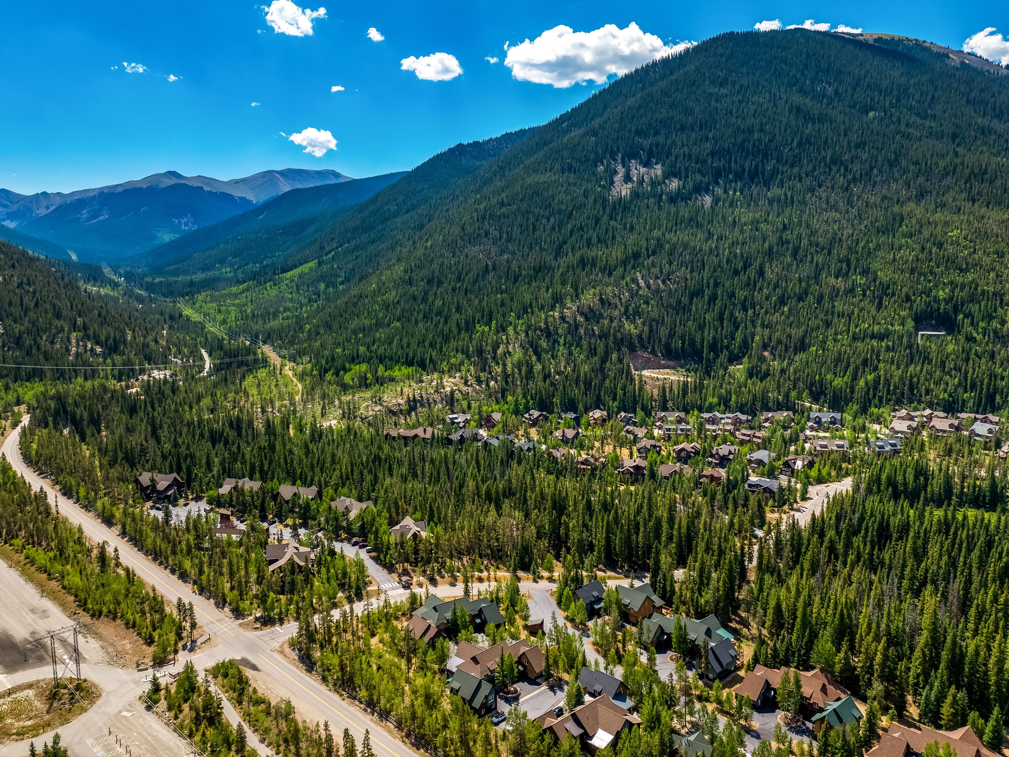 This high-angle aerial view captures a sprawling mountain community nestled within a dense, evergreen forest. The landscape features numerous multi-story residential homes with dark-toned roofs, connected by winding roads and surrounded by towering pine trees. In the background, majestic mountain peaks rise against a vibrant blue sky with scattered clouds, emphasizing the serene and secluded nature of this alpine setting.