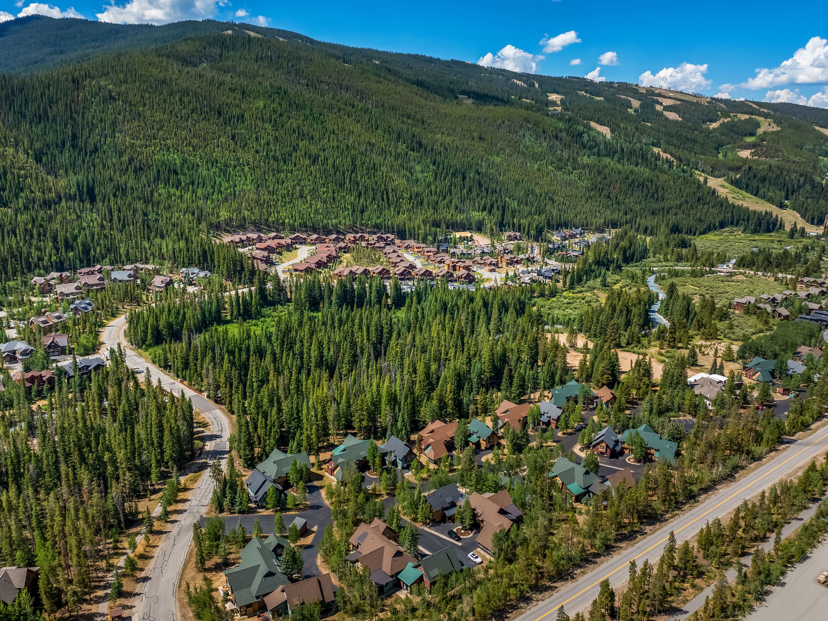 This high-angle aerial perspective captures a sprawling mountain community nestled within a dense, lush evergreen forest. The landscape features numerous detached homes with pitched roofs, winding paved roads, and a scenic stream meandering through the valley floor. The vast, forested mountain slope in the background provides a dramatic and serene backdrop for this residential neighborhood.