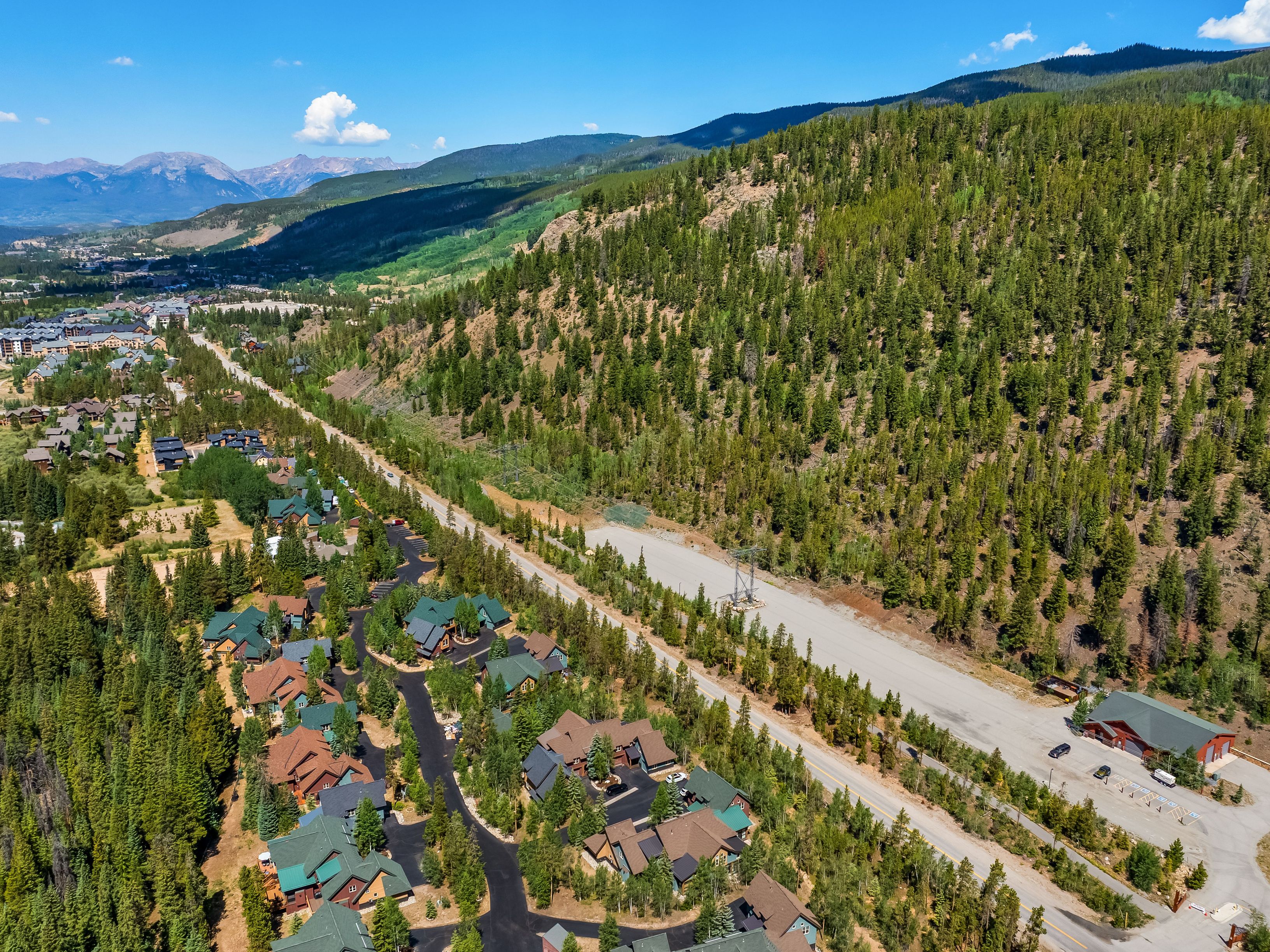 This high-angle aerial perspective captures a mountain residential community nestled at the base of a dense, evergreen-covered hillside. The scene features a collection of multi-story homes with pitched roofs and dark wood siding, connected by winding paved roads, with a clear view of distant, snow-capped mountain peaks under a bright blue sky. A long, straight road runs through the center of the frame, separating the residential area from a large, open gravel lot and a prominent red-roofed building.