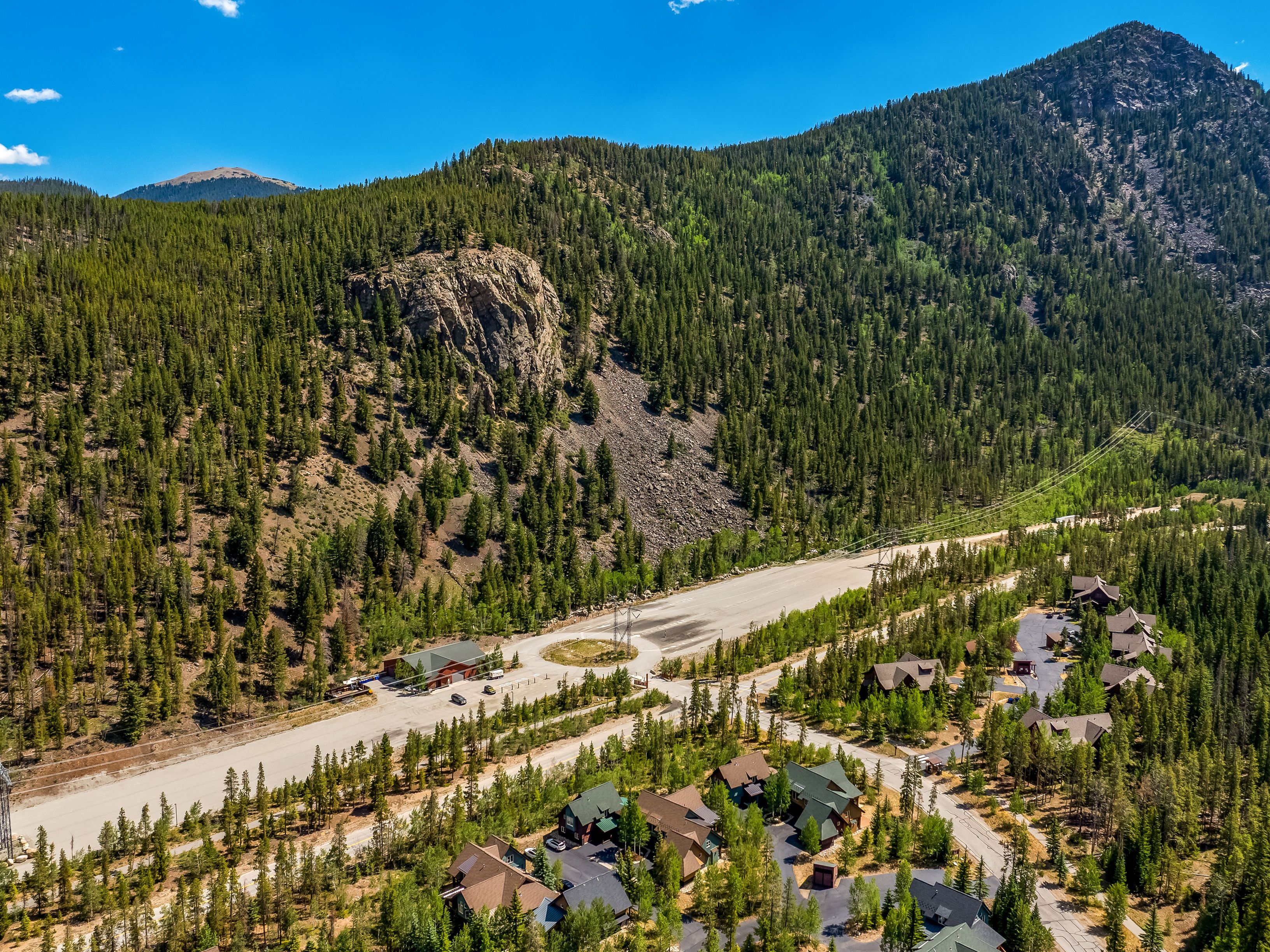 This high-angle aerial view captures a residential mountain community nestled at the base of a vast, pine-covered slope. The scene features a cluster of multi-story homes with dark-toned roofs, a paved circular driveway, and a long, straight road cutting through the landscape. The expansive blue sky and rugged mountain terrain provide a dramatic, scenic backdrop for this secluded neighborhood.