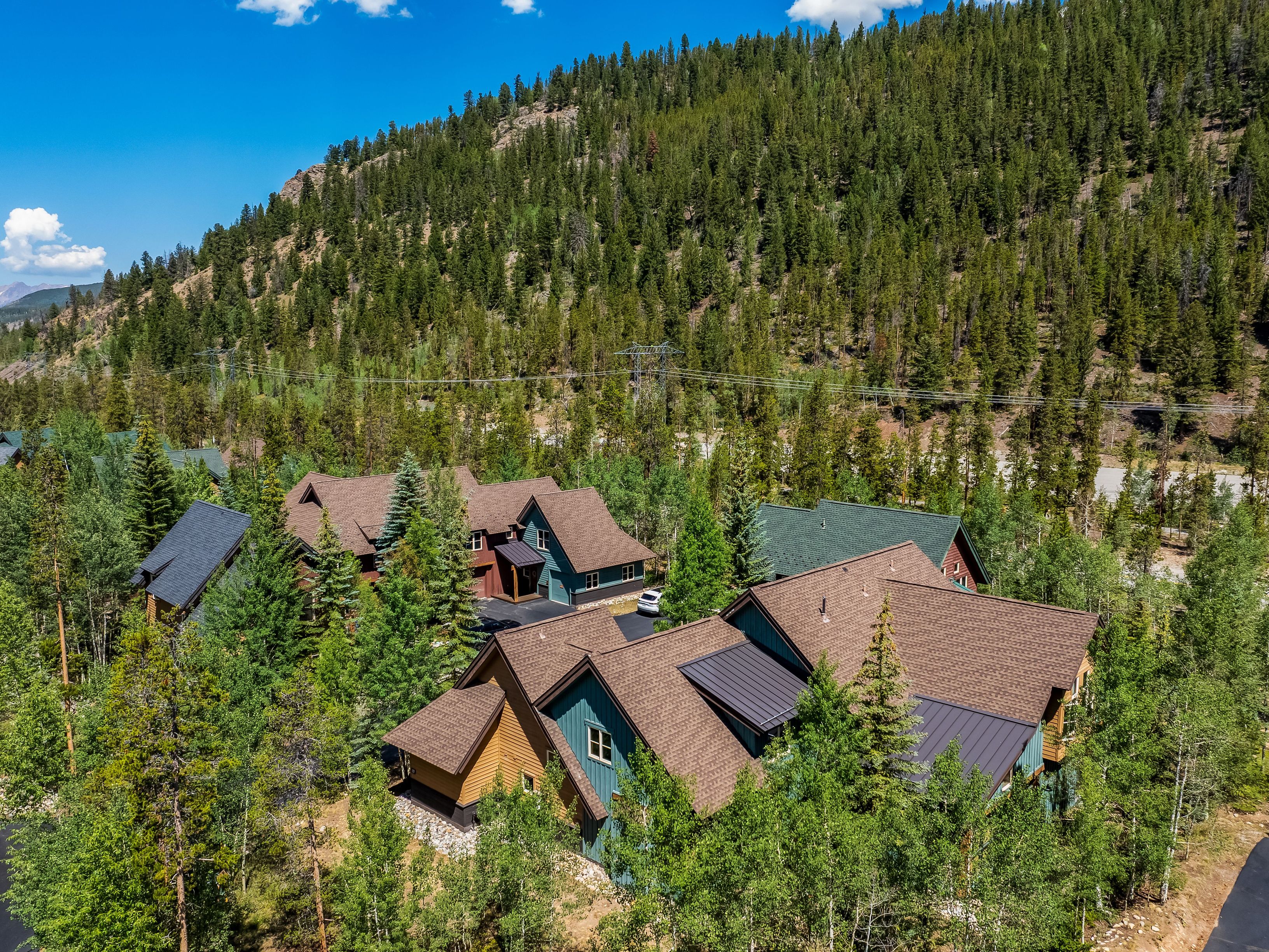 This high-angle aerial view captures a cluster of mountain-style homes nestled within a dense forest of pine and aspen trees. The architecture features multi-gabled roofs with a mix of brown shingles and dark metal roofing, complemented by wood and dark-toned siding. The setting is serene and secluded, with a backdrop of a steep, tree-covered hillside under a bright blue sky.