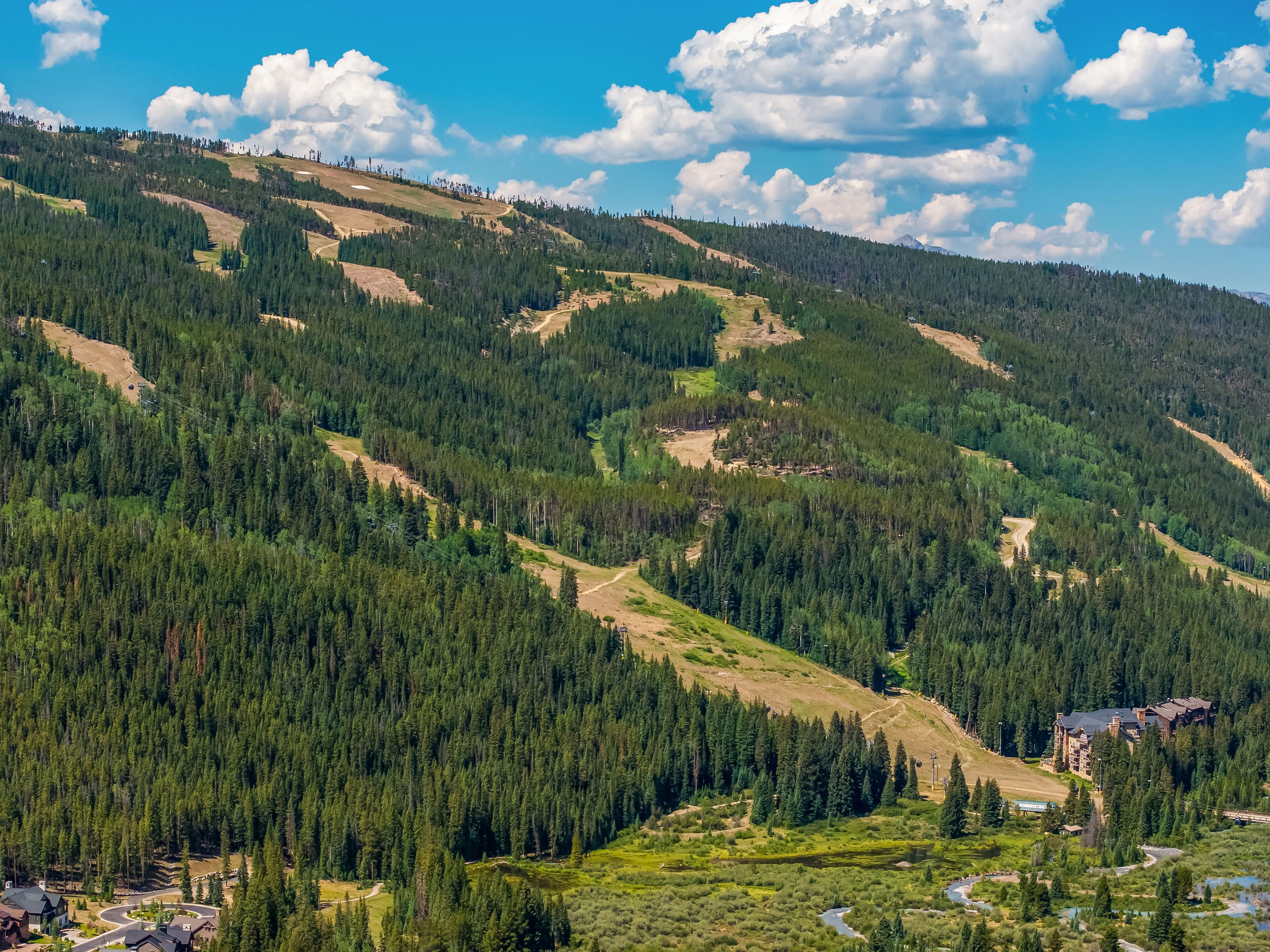 This expansive aerial view captures a mountain landscape featuring a ski resort with cleared trails winding through dense evergreen forests. A large, multi-story lodge with a rustic, timber-and-stone aesthetic is nestled at the base of the slopes near a winding stream and lush meadow. The scene is set under a bright blue sky with scattered white clouds, emphasizing the scenic, recreational nature of the property's location.